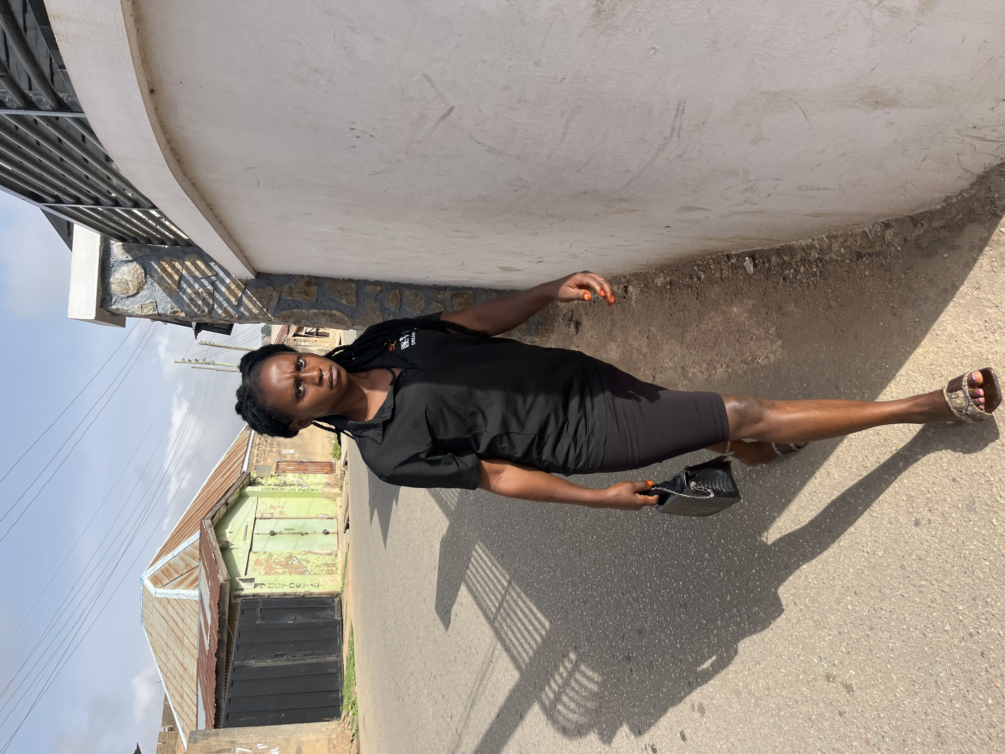 A woman stands on a paved street next to a wall. She is wearing a black dress and sandals and holding a black purse. A building with a corrugated metal roof is visible in the background. The location is likely in Jos, Nigeria, based on the prompt.