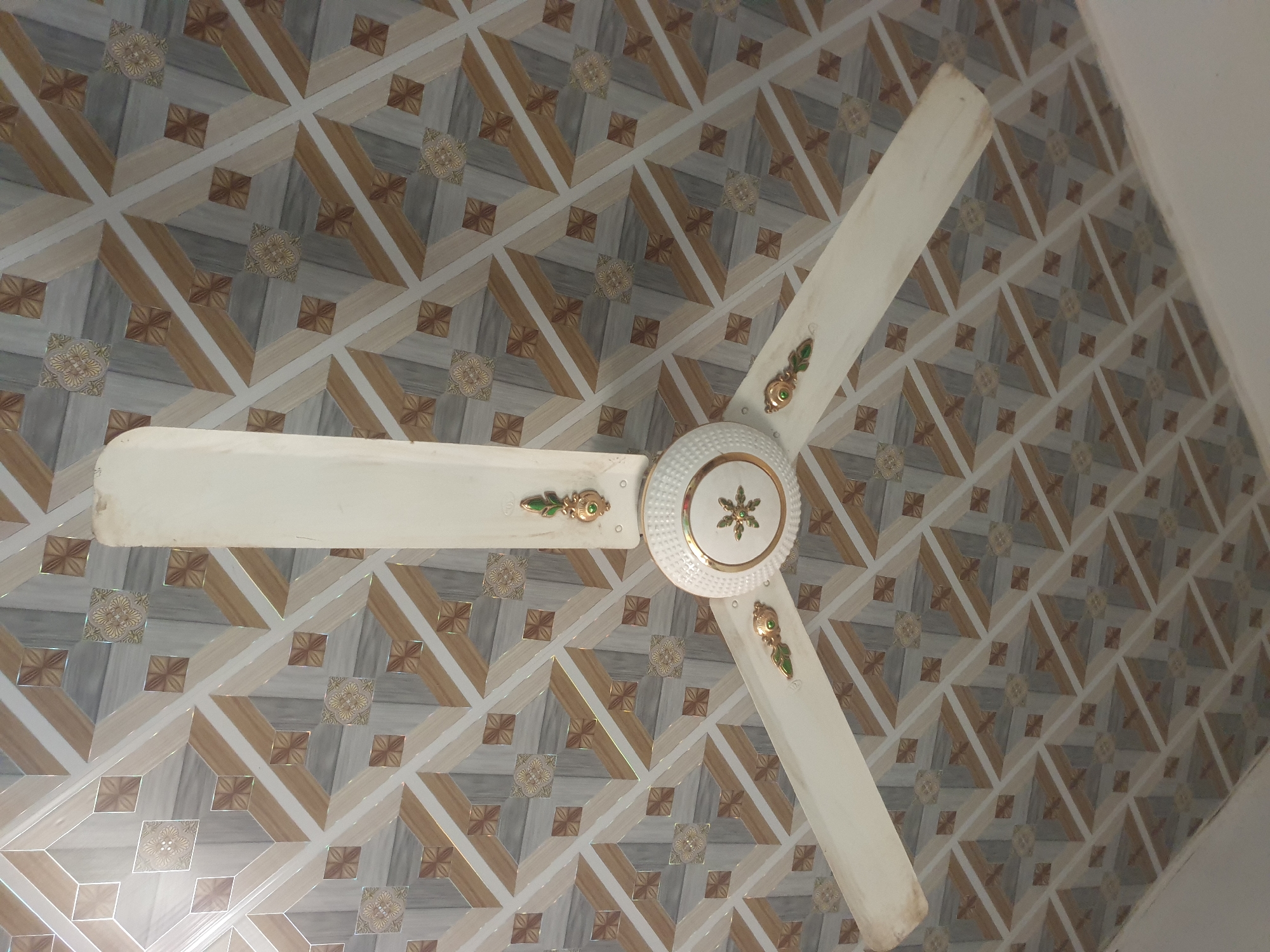 The image shows a three-bladed ceiling fan against a geometrically patterned ceiling in the city of Jalingo, Nigeria. The ceiling features a repeating tessellation of interlocking squares and diagonal stripes in shades of light grey, white, and light brown. Interspersed within this grid are smaller, darker brown square motifs and larger squares adorned with a beige and gold floral-like emblem. The ceiling surface exhibits light reflections, suggesting a glossy finish. The fan is off-white, with its three blades showing slight dust accumulation towards their tips. The central housing is ribbed and white, detailed with a golden ring and a central golden six-pointed star or floral design. Each fan blade is attached via an ornate gold-colored bracket, which incorporates a green oval inlay surrounding a small central gold bead. No people or other significant objects are visible in the scene.