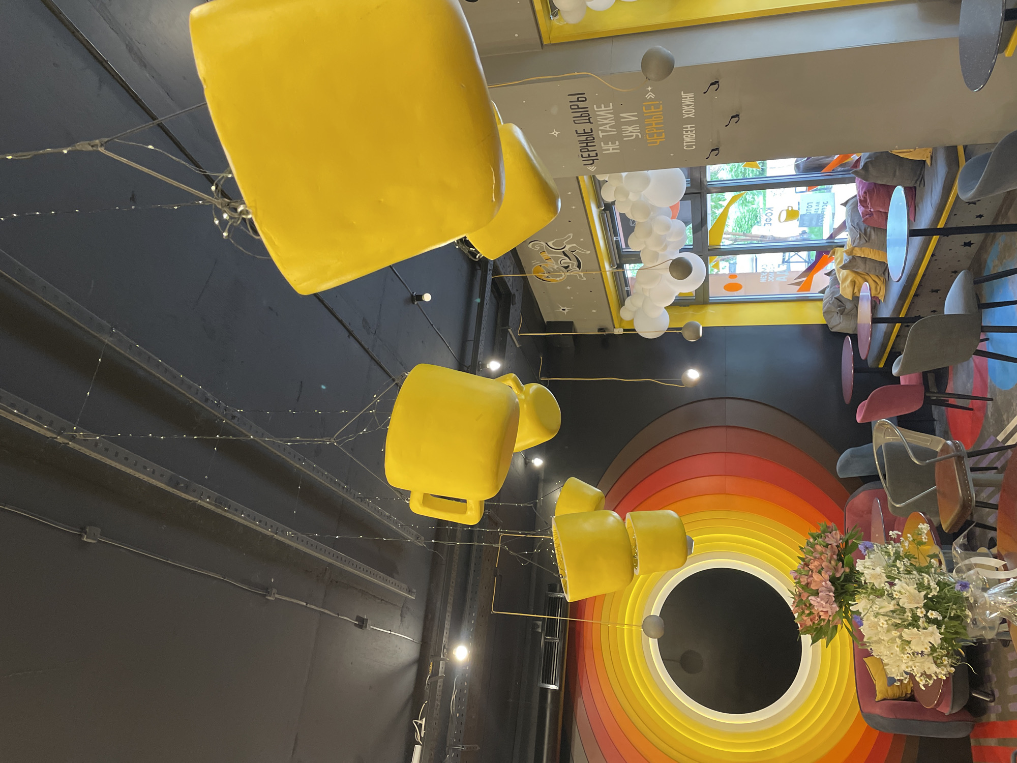 This is an indoor photograph of a cafe interior in Krasnoyarsk, Russia, featuring a black ceiling with exposed conduits and decorative string lights. Several oversized, bright yellow cups are suspended from the ceiling by wires, serving as a quirky decorative element.
On the left side of the image, a large, circular wall art dominates the space. It is a concentric design with a dark center, surrounded by rings of yellow, orange, red, and brown, resembling a stylized black hole or a vibrant sunset. In front of this art, a plush purple sofa with a yellow pillow is visible, alongside a glass-topped table. A vase filled with a bouquet of pink and white flowers sits on a table in the foreground, obscuring part of the sofa.
To the right, a large window offers a view of greenery outside. The window area is furnished with a long, built-in bench seat adorned with colorful throw pillows in yellow, grey, and burnt orange. Small, round tables with dark tops and grey chairs are placed around the seating area. White and grey balloons are clustered near the window, adding to the festive atmosphere.
A gray pillar in the foreground has handwritten text in Russian. The text reads "ЧЁРНЫЕ ДЫРЫ НЕ ТАКИЕ УЖ И ЧЁРНЫЕ! :))" which translates to "Black holes are not that black! :))". Below this, another line reads "СТИВЕН ХОКИНГ" which is "Stephen Hawking". This suggests a thematic or whimsical element to the cafe's decor, likely a reference to the famous astrophysicist.
Various seating options are scattered throughout the cafe, including modern chairs with metal frames and upholstered seats, as well as chairs with dark legs and grey upholstery. The overall ambiance is modern and playful, with a focus on vibrant colors and unusual decorative pieces. The lighting appears to be a mix of ambient string lights and overhead spotlights. No people are visible in the frame.