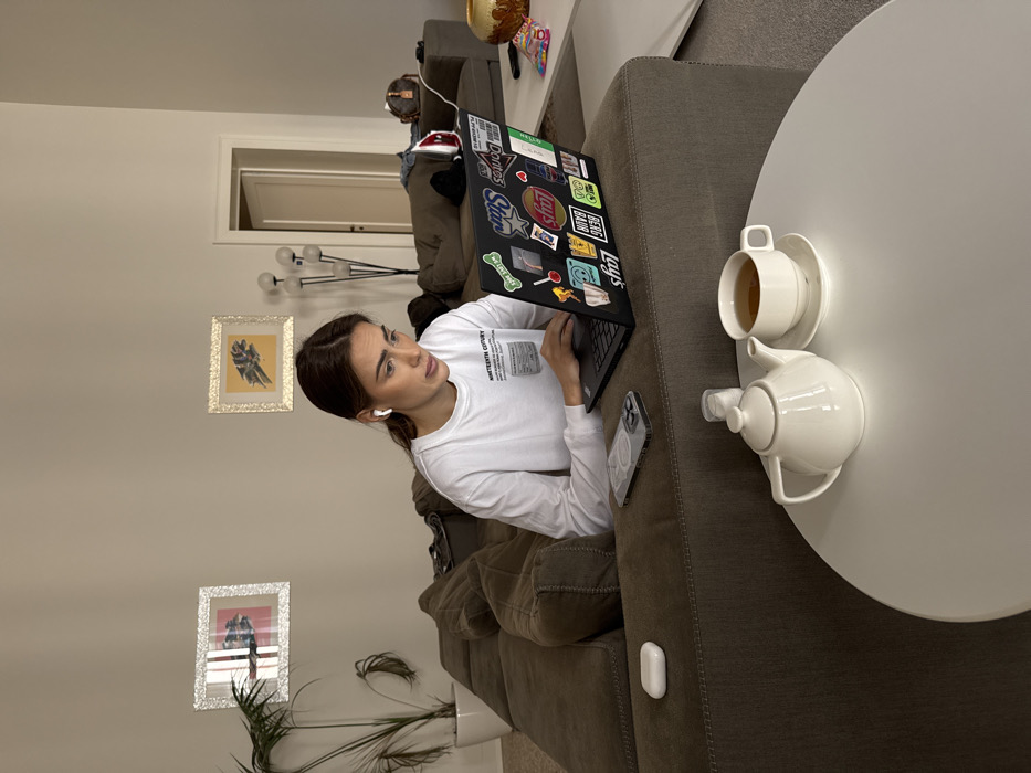 The scene depicts an interior residential setting, identified as being located in Venice, Italy. A young adult female, with dark hair pulled back and wearing a white long-sleeved t-shirt, is seated on a large brown sectional sofa, actively engaged with a black laptop computer. She wears white wireless earbuds in both ears and maintains a focused expression while typing on the laptop's keyboard. The laptop lid is adorned with numerous colorful stickers, including brand logos like "Lay's" and "Doritos."
On the sofa cushion to her left, a clear smartphone case and a white wireless earbud charging case are visible. In the immediate foreground, on a white round table, a white ceramic teapot, a white teacup filled with amber liquid on a matching saucer, and a small white shaker are arranged.
The background shows light-colored walls adorned with two framed art pieces: one with a silver, textured frame and an abstract image featuring red elements, and another with a gold, textured frame containing an image with a yellow background and a dark, organic shape. A tall potted plant in a white container stands in the left corner. To the right, behind the sofa, a floor lamp with multiple spherical lights is present. A white doorframe leads to an adjacent room. Further down the sofa, a brown duffel bag and a red-and-white iron are resting. A golden-brown decorative vase and several small, colorful candy packets are visible on a low white surface to the far right. The floor appears carpeted in a light neutral tone.