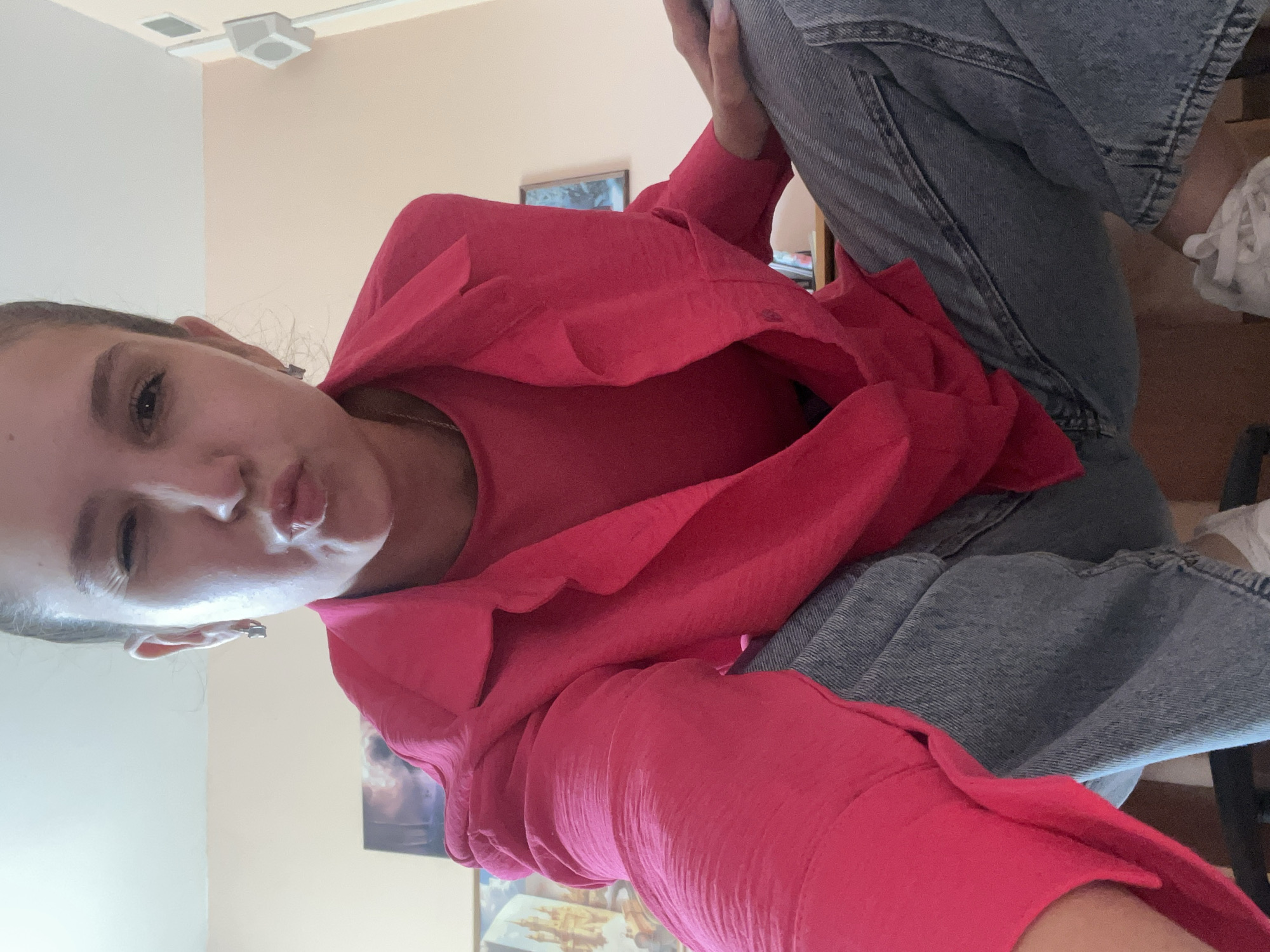 The image shows a young woman with her hair pulled back, wearing a bright pink collared shirt and blue jeans. She is making a kissing face and winking at the camera. The background appears to be an indoor setting, possibly a room with light-colored walls and some framed artwork on the wall. The camera angle is from a low perspective, looking up at the woman.