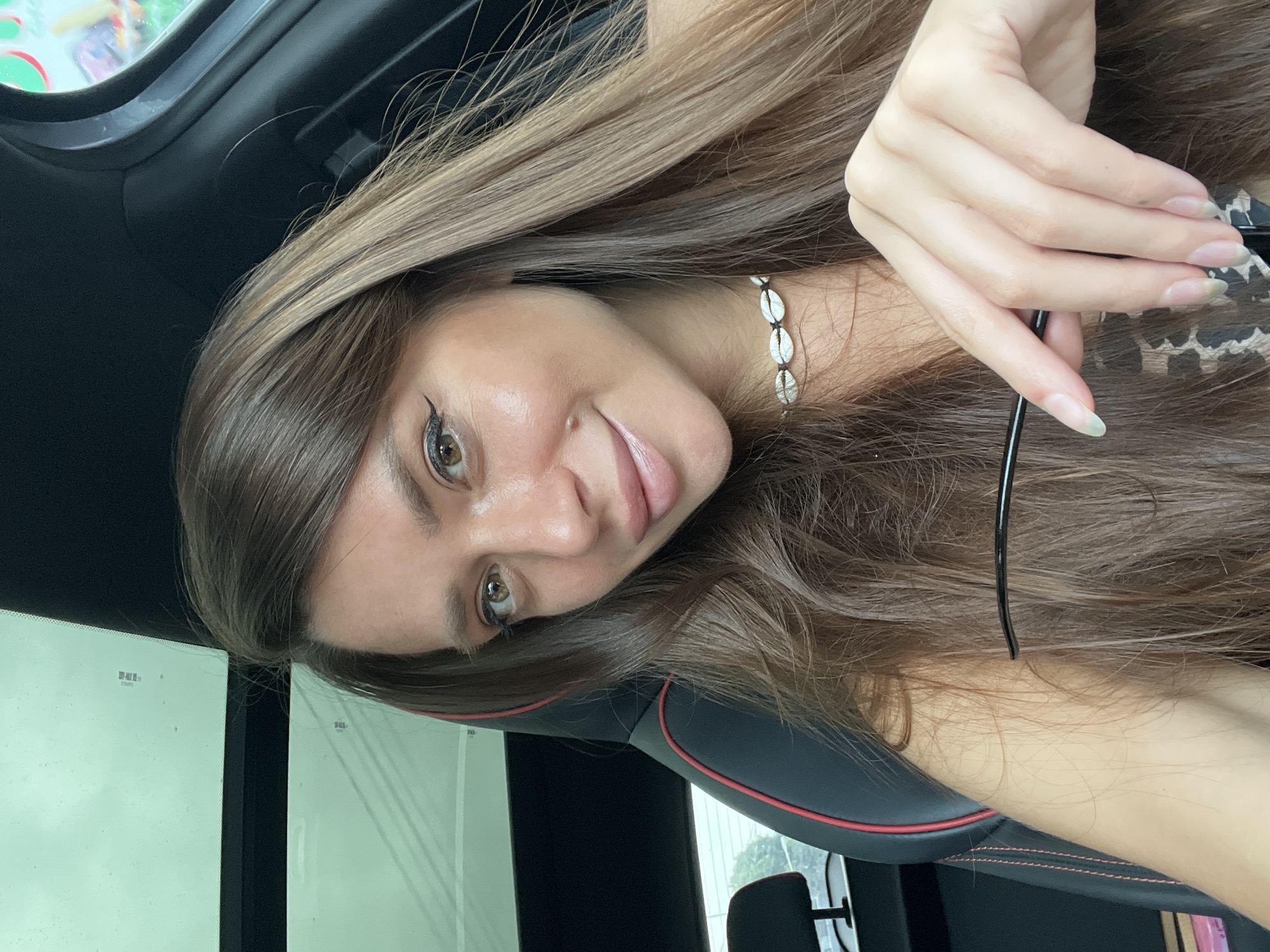 A rotated close-up portrait shows a young woman with long, straight brown hair, possibly taken inside a car. Her head is tilted, and she gazes directly at the viewer with a gentle smile, revealing dark eyes with prominent winged eyeliner. She wears a necklace made of white cowrie shells. Her hand, with natural-looking nails, holds a pair of black-framed glasses.
The setting is an automobile interior, evidenced by the dark car seat with red accent stitching and a car door frame. Bright daylight streams in through the window, suggesting it is daytime. Faint text, including "50%-RUV" and some indistinct Thai characters or symbols "บปส.", is visible on the window glass. The woman's expression conveys a sense of calm and pleasantness, indicative of a relaxed, casual moment. This scene is contextualized to Baan Hua Thanon, Thailand.