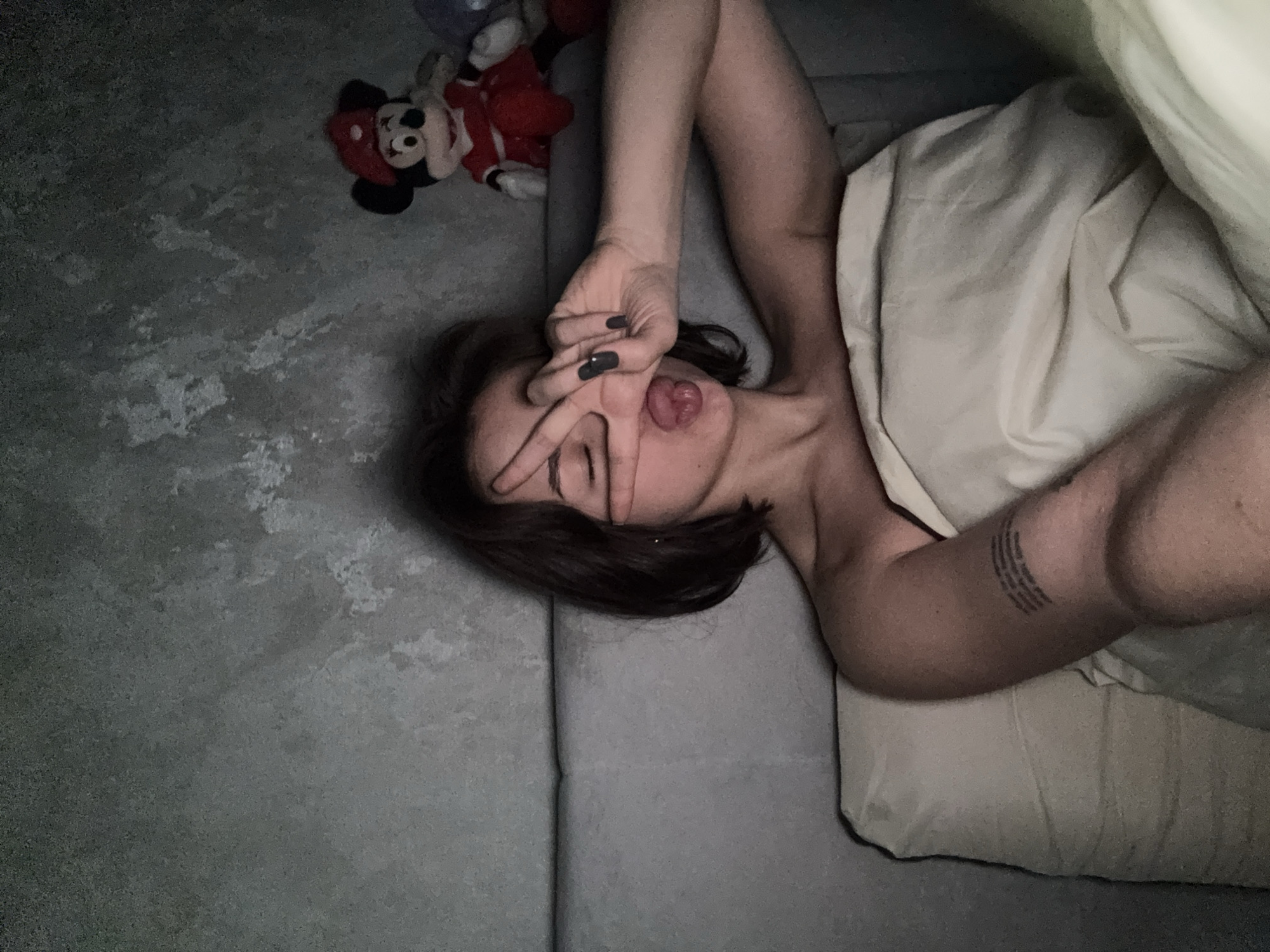 A person is captured in a playful, relaxed pose indoors, likely in Лобаново, Russia. The individual, appearing to be female, lies diagonally across the frame on a grey upholstered surface, possibly a bed or couch. Their left hand, adorned with dark nail polish, makes a peace sign over their face, partially obscuring their eyes, while their tongue is playfully sticking out. Dark brown hair frames their face, and a multi-line text tattoo is visible on their left upper arm.
A light-colored duvet or blanket covers part of their body. To the upper left, on a textured grey surface (possibly a floor), two stuffed toys are visible, including a prominent Mickey Mouse character in a red outfit. The lighting is dim, suggesting a nighttime or low-light setting. The overall mood conveyed is one of casual comfort and playfulness.