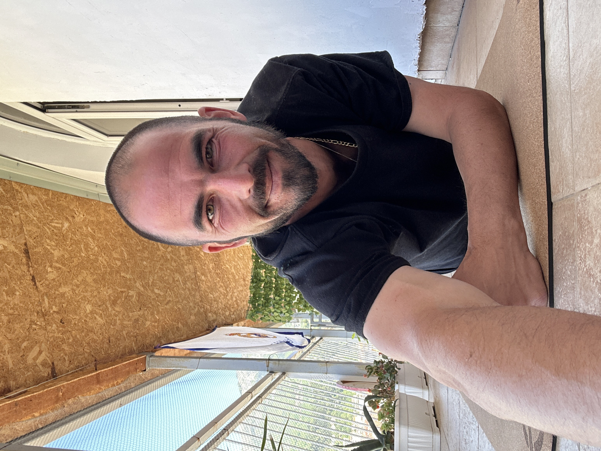 A man with a shaved head, dark beard, and wearing a black t-shirt and gold necklace is taking a selfie. He is smiling slightly. The background shows a partially enclosed balcony or patio with a metal frame and wire mesh. There is a white Real Madrid jersey hanging on the balcony. There are potted plants with greenery visible outside the mesh. The floor appears to be tiled. Some architectural details suggest this is a building in a coastal area. Given the context, it is possible this photo was taken in Budva, Montenegro, but this cannot be definitively confirmed from the image alone.