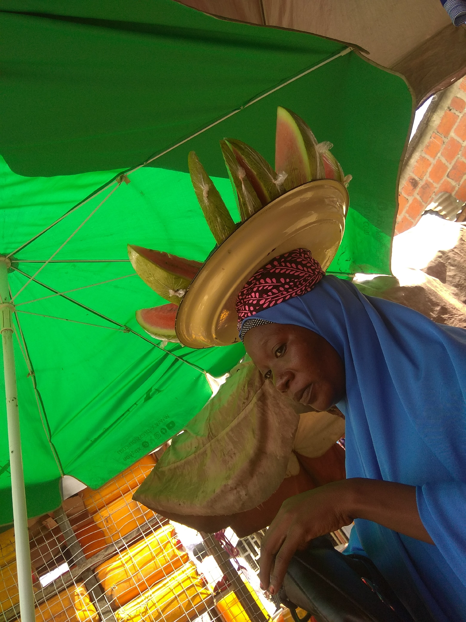 A woman wearing a blue hijab and a patterned headscarf is the central subject. She is looking down and to her left, with a neutral expression. She is carrying a large, golden bowl on her head, filled with slices of watermelon. The watermelon slices are wrapped in plastic.
The scene is outdoors, likely a market, indicated by the presence of an umbrella providing shade. The umbrella is green and the structure supporting it is visible. Behind the woman, a brick wall and a corrugated metal roof are visible, suggesting a semi-permanent or informal structure. In the foreground, below the woman and to her left, a large, beige, leaf-like object is partially visible, as well as some yellow objects stacked behind a wire mesh.
The lighting suggests it is daytime, with sunlight filtering through the umbrella. The overall impression is of a street vendor selling fruit. The location context of Jalingo, Nigeria, aligns with the visual cues of an outdoor market setting. No specific text is clearly legible in the image, though there appears to be some faint printing on the umbrella.