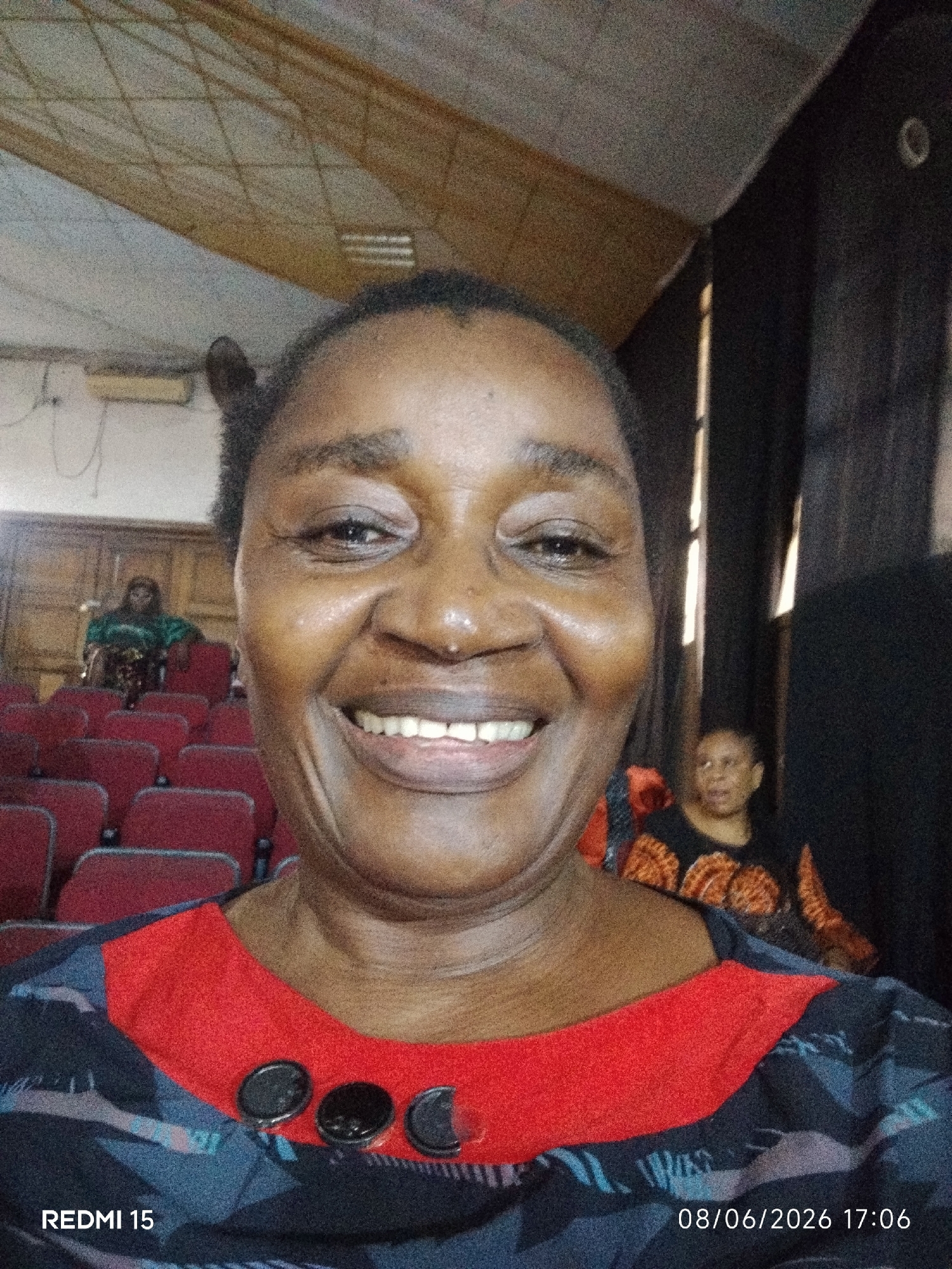A medium shot captures a smiling woman in the foreground, her face filling most of the frame. She is wearing a dark top with a red neckline and three black circular buttons. Behind her, in the mid-ground, are rows of red theater-style seats. Further back, two other women are visible, one sitting in a seat and another standing. The environment appears to be an indoor venue, possibly an auditorium or conference hall. The image includes text overlays indicating "REDMI 15" in the lower left corner and a date and time stamp "08/06/2026 17:06" in the lower right corner. The ceiling features exposed wooden beams and is lit by fluorescent lights. The overall lighting is somewhat dim. The location is identified as Aba, Nigeria.