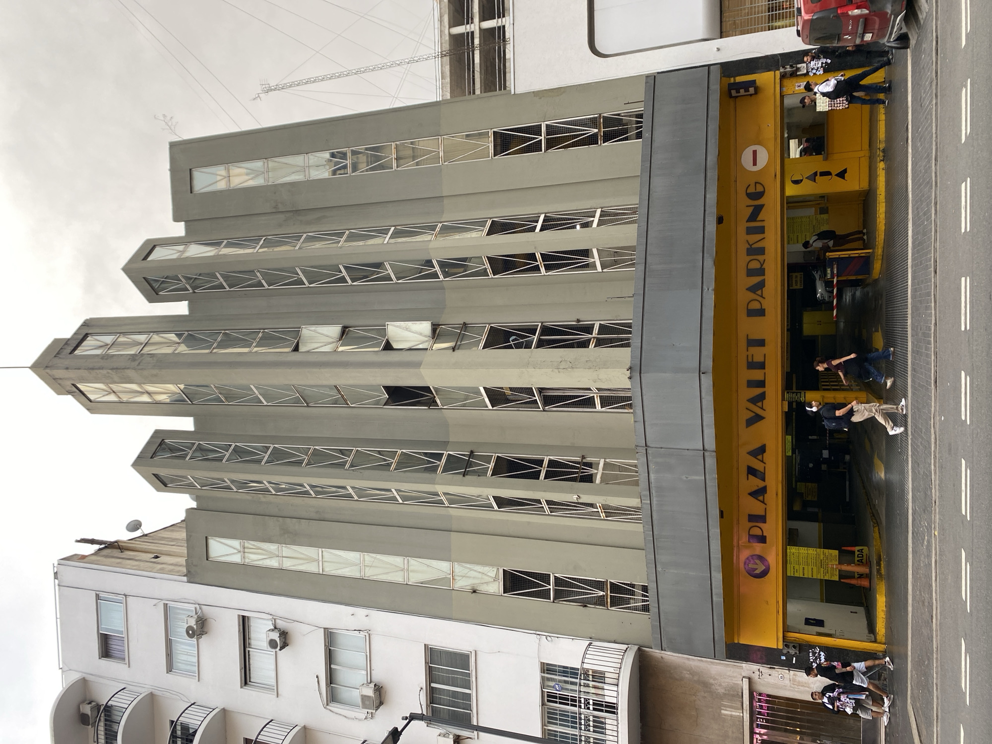 The image depicts the street-level entrance and multi-story facade of a parking garage identified as "PLAZA VALET PARKING" at Carlos Pellegrini 1025, Buenos Aires, Argentina. The prominent central building features a distinctive Art Deco-style architectural design, characterized by a pale greenish-grey concrete facade with multiple vertical, recessed window columns composed of small, geometric glass panes within light-colored frames. Several upper windows are open, tilted outwards.
The ground floor, covered by a dark grey overhang, presents a bright yellow facade with the purple text "PLAZA VALET PARKING" and a downward arrow to the left, and a "no entry" sign to the right. Two vehicle entrance/exit lanes are visible below, separated by a concrete median. A red and white barrier arm is raised in the right lane. Inside the entrance, yellow informational signs, orange traffic cones, and a "CAJA" (cashier) sign are present.
To the immediate left of the parking garage, a white multi-story residential or commercial building with several balconies, black metal railings, and visible exterior air conditioning units is present. Its lower levels feature barred windows. To the right, a portion of another white building is visible. In the upper right background, a tall, slender metal lattice tower structure rises against an overcast sky.
Approximately 10-12 individuals are present on the sidewalk and around the parking entrance. On the left sidewalk, three males are walking, two in dark t-shirts and light shorts. Near the center, a person wearing headphones and a backpack walks rightwards. On the right side, individuals are seen entering or exiting the parking facility, including a person carrying a box near a partially visible red panel van parked at the curb. The street is asphalt with white dashed lines and appears wet, indicating damp conditions under a cloudy sky. A light-colored vehicle is partially visible inside the parking structure.