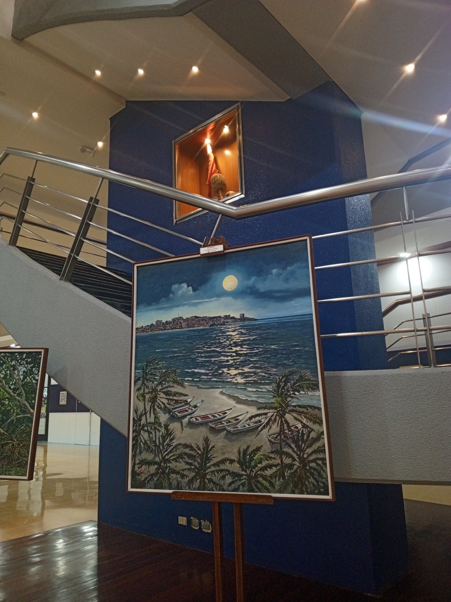 The image depicts an indoor gallery or museum space, likely in Porlamar, Venezuela. A large painting dominates the foreground, depicting a beach scene at night with palm trees and boats under a full moon. The building in the background of the painting is suggestive of the skyline of Porlamar. The artwork is displayed on an easel in front of a dark blue structural column. To the left, a smaller painting is visible in the periphery. The building features a staircase with metal railings. Above the main painting, set into the column, is a display case containing a statue or artifact. The architecture includes a mix of modern materials and design. The floor is polished, reflecting the light.