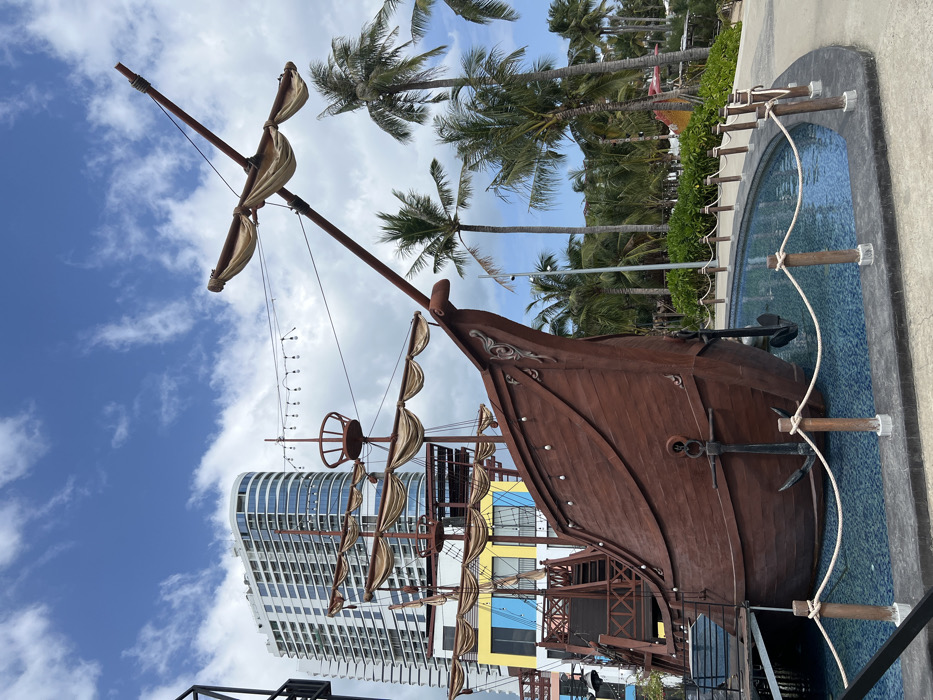 A brown wooden pirate ship model stands prominently against a backdrop of a modern white high-rise building and a blue, cloudy sky. The ship's mast extends upward, adorned with tan sails and ropes. Palm trees line the background, adding a tropical feel. A small, rectangular water feature surrounds part of the ship's hull, secured by rope and wooden posts. The model is partially elevated.