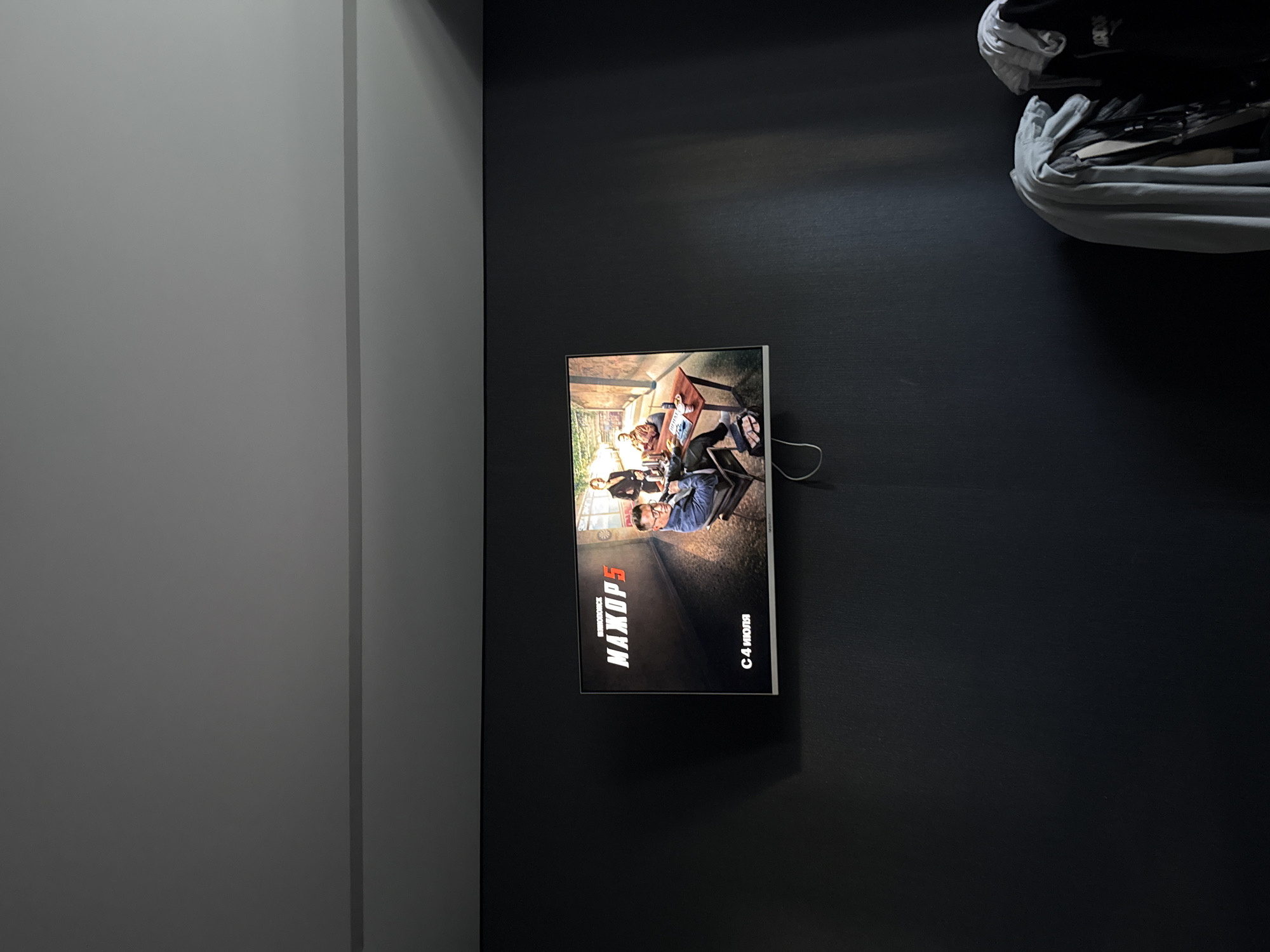 The image shows a modern interior space, likely a living room or entertainment area, with a dark, textured wall. A flat-screen television is mounted on the wall, displaying a movie poster or title screen. The screen features a title in large, bold, stylized Russian letters that read "МАХОР 5", with smaller text above it that appears to be "БОГАТИР". Below the title, text reads "С 4 ИЮЛЯ", indicating a release date of July 4th. The screen also shows an image of several people, possibly actors, seated around a table in an outdoor or semi-outdoor setting, with a hint of greenery in the background.
In the upper right corner of the image, a large, light-colored bundle of fabric, possibly a blanket or duvet, is draped over a surface, partially obscuring a black object, which might be a bag or backpack. The lighting in the room suggests it is daytime, with light casting shadows on the wall from an unseen source, possibly a window or lamp. The overall mood is contemporary and minimalist, with the television and its display serving as the focal point. The location context provided is Velikiye Luki, Russia, which is consistent with the Russian text displayed on the screen.