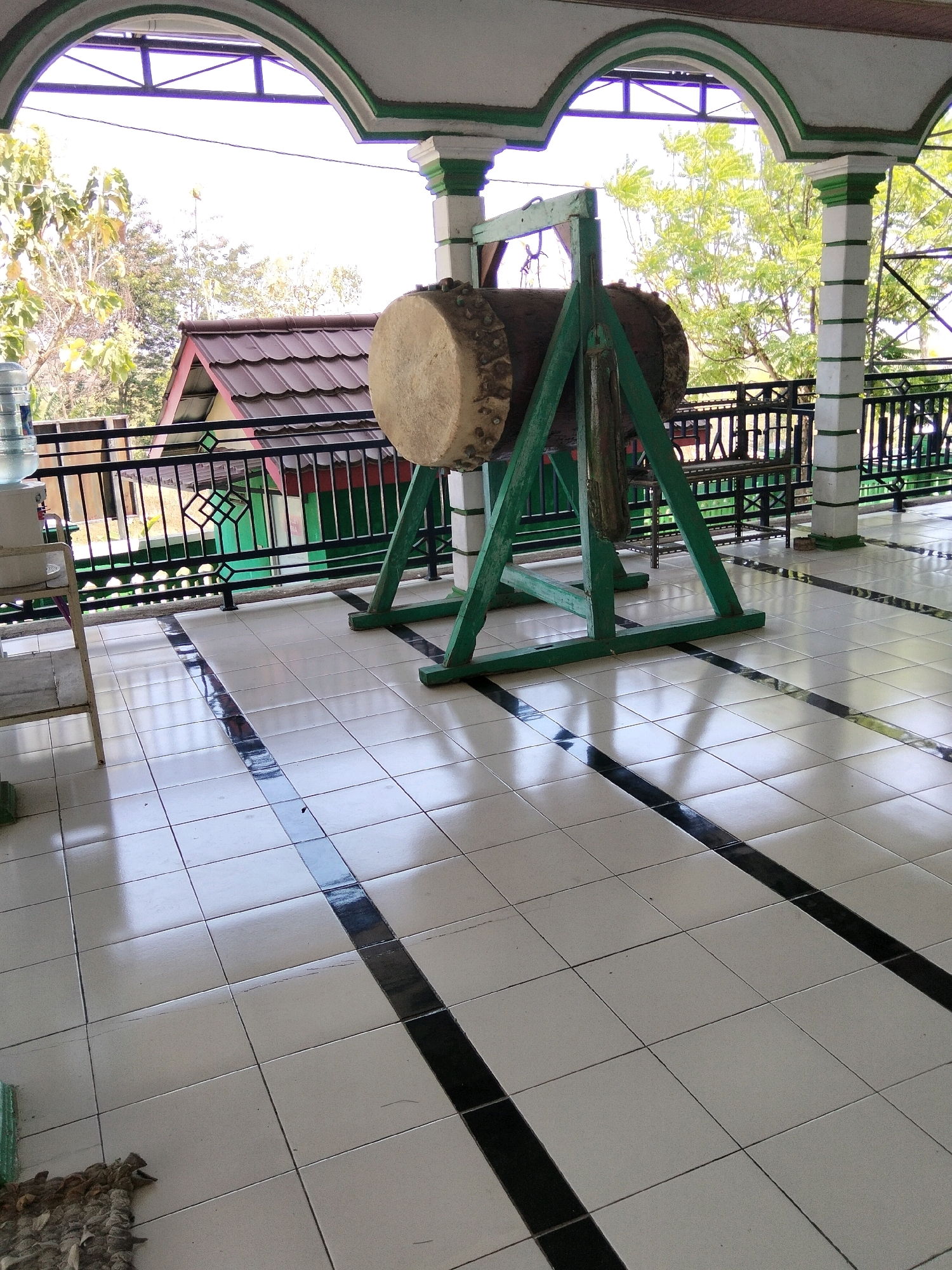 An outdoor shot captures a large wooden drum, known as a "bedug," situated on a green triangular stand. The drum is positioned on a white tiled floor with black accent stripes, reflecting the bright daylight. Behind the drum, a dark metal railing encloses a porch or balcony area. Beyond the railing, trees with lush green foliage are visible, along with a red-tiled roof of a building. The scene appears to be in Jenangan, Indonesia.
