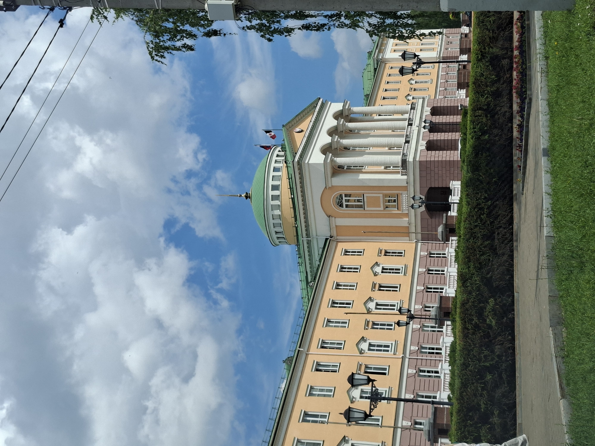 The image shows the Udmurt State University in Izhevsk, Russia, on a cloudy day. The university building is a large, neoclassical structure with a prominent dome and a colonnade. The building is painted in a pale yellow color with white trim. The dome is green and topped with a spire. Two flags fly from the roof of the central portico, one appearing to be the flag of the Udmurt Republic and the other the Russian flag. In front of the building is a neatly trimmed hedge. Lampposts are visible along the walkway. In the foreground, there is a paved area with grass and flower beds. A utility pole with wires and a street light fixture is visible on the right side of the image, with tree branches obscuring part of the sky. The sky is blue with large, white cumulus clouds.