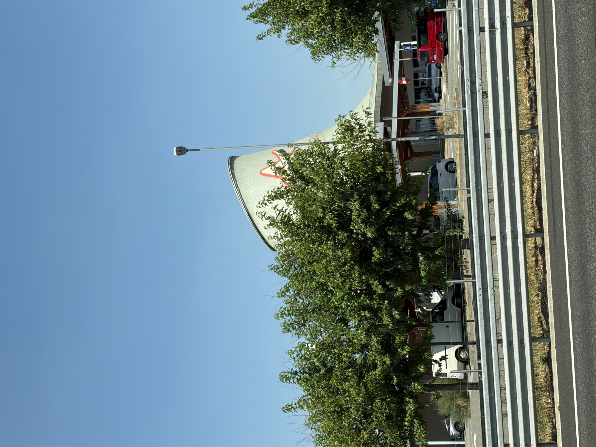 The image shows a view from a highway of a modern building with a curved, silver roof that has a large red logo on it. In front of the building, there is a parking area with several vehicles, including a white van, a silver car, and a red minivan. Trees and landscaping are visible in the foreground and around the building. A street light is also visible in the sky. The sky is clear and blue. The building appears to be a commercial establishment, possibly a large retail store or a service center. Based on the style of architecture and signage, this building is likely located in Lainate, Italy, as indicated by the context. The scene is captured on a sunny day.