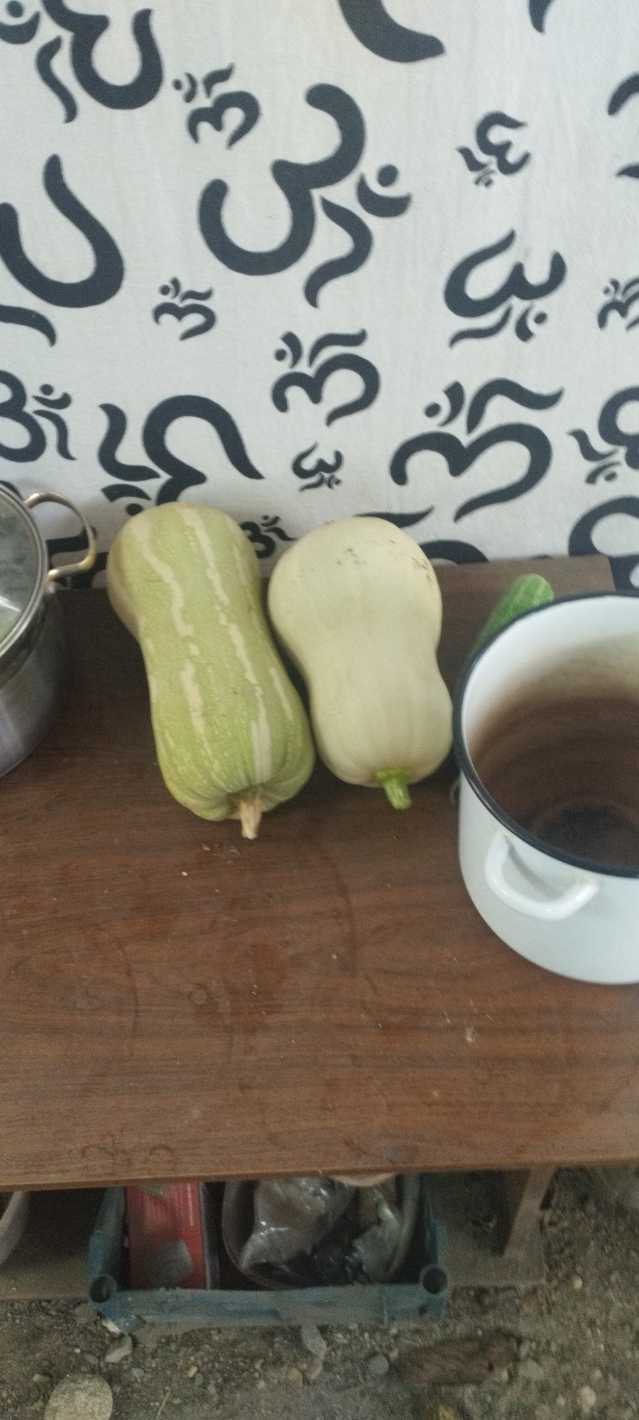 The image shows two light-colored gourds, one with green stripes, resting on a wooden surface. To the left, a metal pot with a handle is partially visible. To the right, a white enamel pot with dark trim is positioned. In the background, a white fabric is adorned with a repeating pattern of dark grey "Om" symbols. Below the wooden surface, a black metal rack holds various items, including what appears to be a red box and plastic bags filled with material, situated on a gravelly ground. The scene is indoors or under a covered area, with a backdrop suggesting a wall or hanging textile. The lighting is diffused, and the overall composition is a close-up view of the gourds and surrounding objects. No specific geographical markers indicate Yaroslavskaya, Russia.