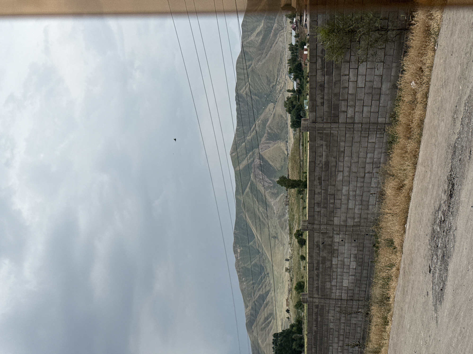 The image depicts a landscape scene, likely in a rural or mountainous area. The primary subjects are a mountain range with visible terracing or erosion patterns, a brick wall, a road, and a cloudy sky.
The mountain range dominates the background, appearing green and somewhat barren, with clear lines indicating topographical features. A small settlement with buildings can be seen nestled at the base of the mountains. Power lines stretch across the sky in the middle ground, adding a man-made element to the natural landscape.
In the foreground, a grey brick wall runs horizontally across the image, separating the viewer from the scene beyond. To the right of the wall is a paved road with some patches of dark debris or asphalt. Between the wall and the road, dry, yellow grass grows.
The sky is overcast with thick, grey clouds, suggesting a dull or potentially rainy day. There is no activity or event explicitly taking place; it appears to be a still, observational shot. The lighting is diffused, consistent with cloudy weather. The overall mood is neutral, leaning towards somber due to the overcast sky. No visible text is present in the image. The orientation of the image is vertical, with the top of the frame appearing to the left.