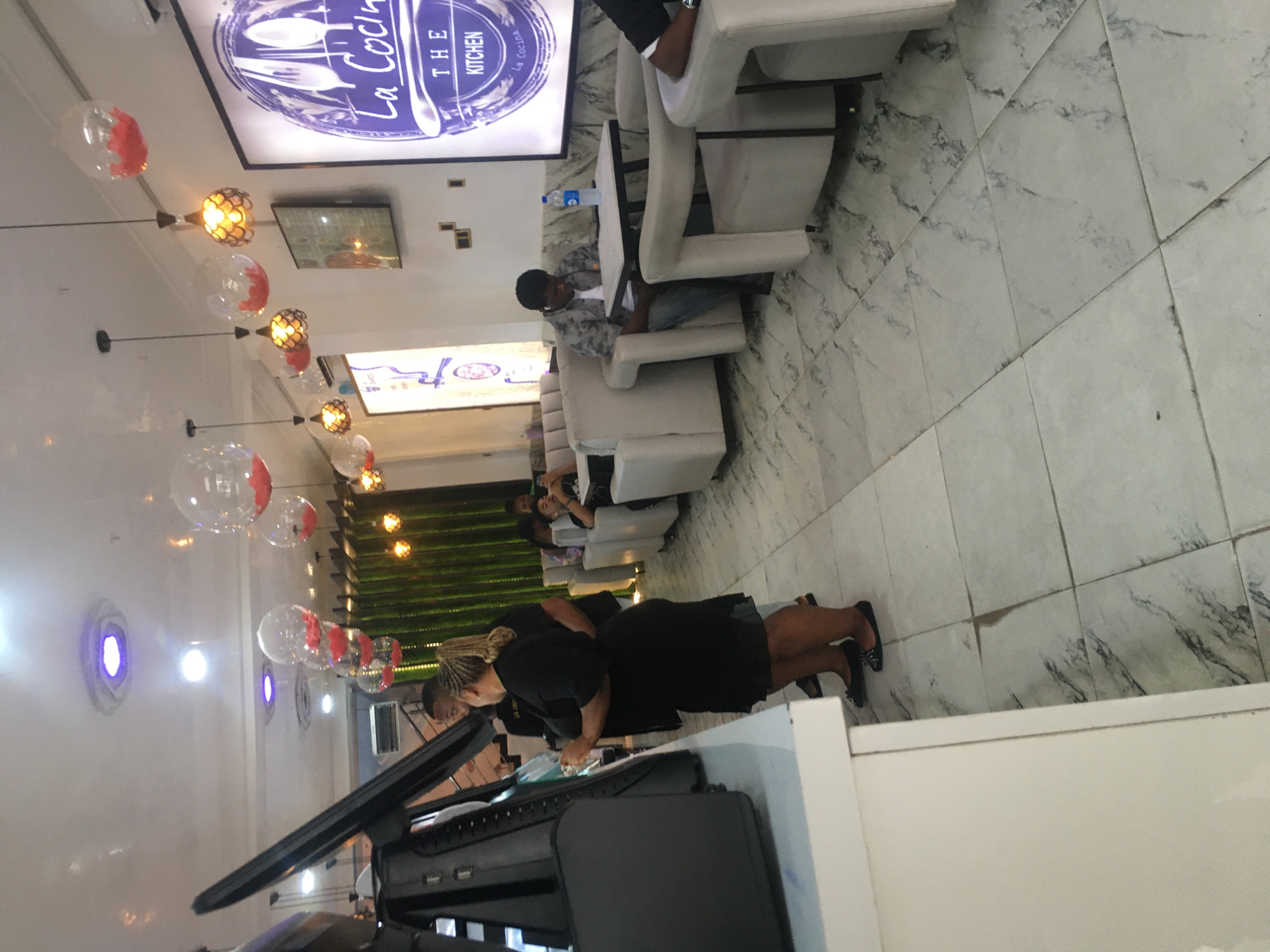 The image depicts the interior of "La Cocina: The Kitchen," located in Ado, Nigeria. A waitress in a black dress stands at a counter, likely preparing orders or tending to the point of sale system. Several patrons are seated in white upholstered chairs at tables, one of whom appears to be looking at a mobile phone. There are decorative balloons and light fixtures on the ceiling, and green foliage is visible in the background. The floor is tiled.