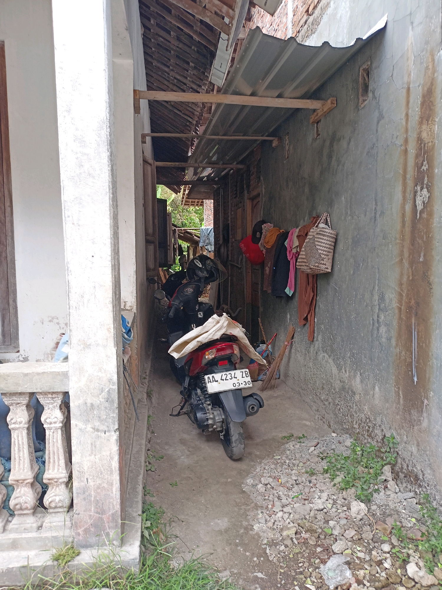 The image depicts a narrow outdoor walkway between two buildings in Ngepanrejo, Indonesia. The walkway has a concrete floor and is partially covered by a corrugated metal roof supported by wooden beams, suggesting it is an alleyway or a service entrance.
On the left side, a white concrete pillar with decorative molding is visible, part of a structure with a wooden frame. On the right, a rough, weathered concrete wall displays water stains and cracks. Hanging on this wall are several items of clothing (a red cap, a brown garment, a pink garment, a dark garment, and a light-colored garment) and a woven basket. A broom leans against the wall near the ground.
Two motorcycles are parked in the walkway. The one in the foreground is red and black, with its rear visible. It has a license plate that reads "AA 4234 ZB 03-30". A helmet rests on the handlebars of the motorcycle behind it, which appears to be black. The area around the motorcycles is cluttered with debris, including rocks, dirt, and some plastic waste. Small green plants are growing amongst the debris. The overall impression is one of a utilitarian, somewhat neglected outdoor space.