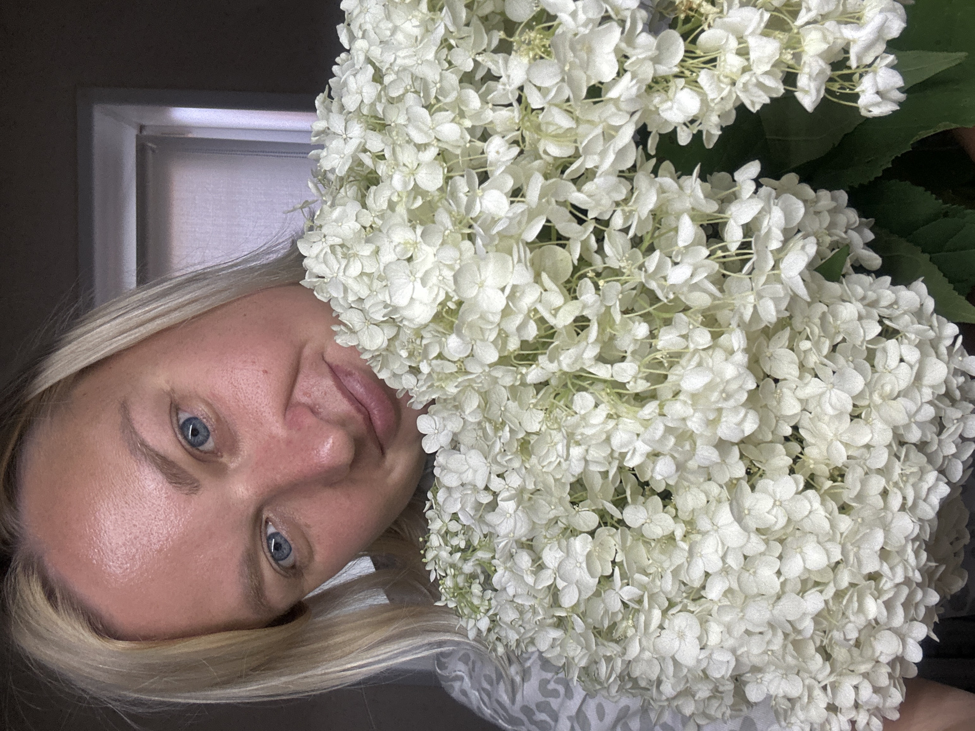 A blonde woman with blue eyes is holding a large bouquet of white hydrangeas in front of her face. She is looking directly at the camera with a slight smile. The background appears to be indoors, possibly in a home, with a window visible in the upper right corner. The image does not contain any information about the city of Chaikovsky, Russia, or any other specific location.