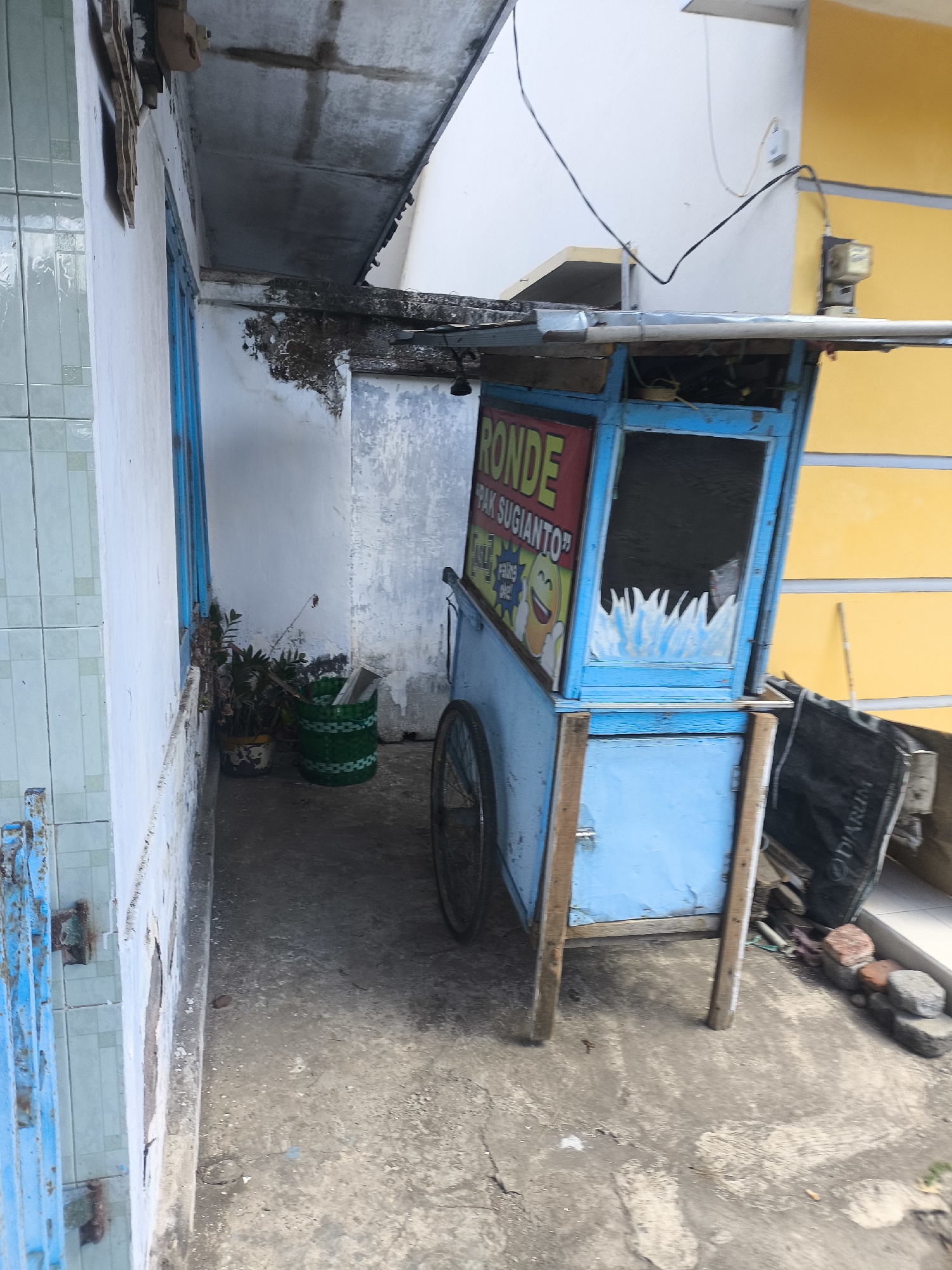 The image depicts a street scene in Lumajang, Indonesia. A bright blue food cart, possibly for selling "Ronde Pak Sugianto" as indicated by the signage, is prominently featured. The cart has a glass display area with what appear to be decorative white elements inside. It is supported by wooden legs and has a wheel visible on the right side. The cart is parked on a concrete ground, with a yellow wall and a white wall in the background. To the left of the cart, there is a white tiled wall and a blue door or gate. In front of the white wall, there are two potted plants, one with dark green leaves and the other a green woven basket containing some items. The environment appears to be an outdoor alleyway or a narrow street with some debris on the ground.