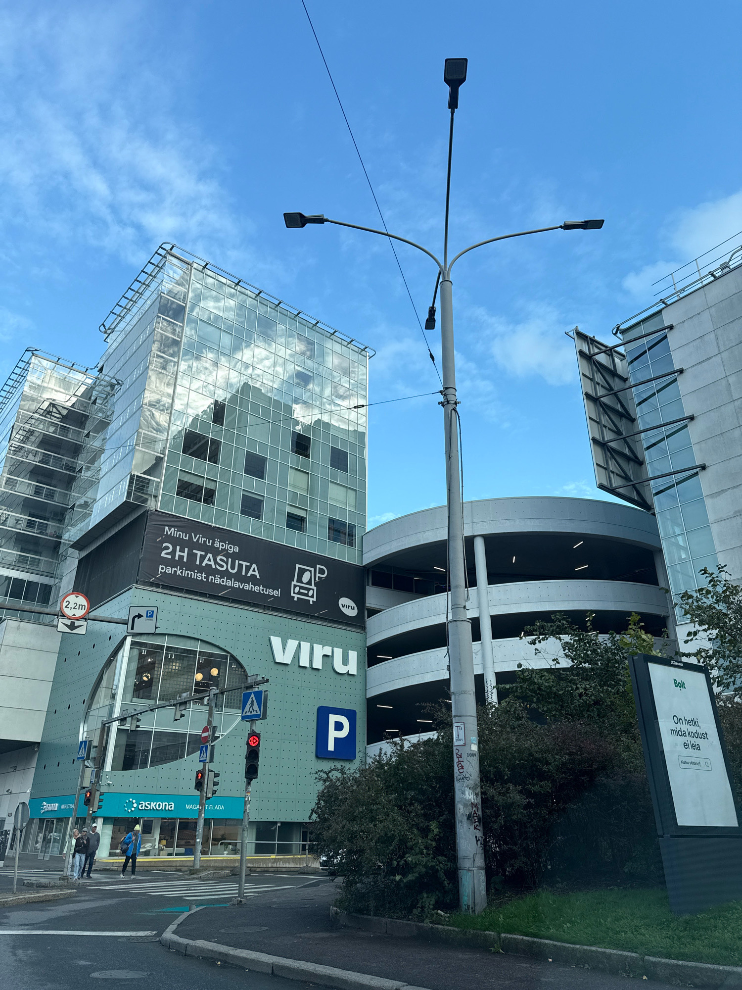 A street-level, medium shot captures a section of the Viru Keskus shopping center in Tallinn, Estonia, on a cloudy day. The foreground shows a paved road with a pedestrian crossing. In the midground, a modern, multi-story building dominates the scene with a glass facade reflecting the sky. A large banner displays "Minu Viru äpiga 2H TASUTA parkimist nädalavahetusel" (With my Viru app, 2 hours free parking on weekends), featuring a car and parking symbol. Below this, a green facade of the shopping center is visible with the "Viru" logo. Traffic lights show red, and pedestrians are crossing the street. A lamppost with multiple lights stands in the center, with wires extending upwards. To the right, a multi-level parking garage with a curved structure is visible, partially obscured by trees. A digital advertisement board displaying "Bolt" and Estonian text is present on the right side of the frame.