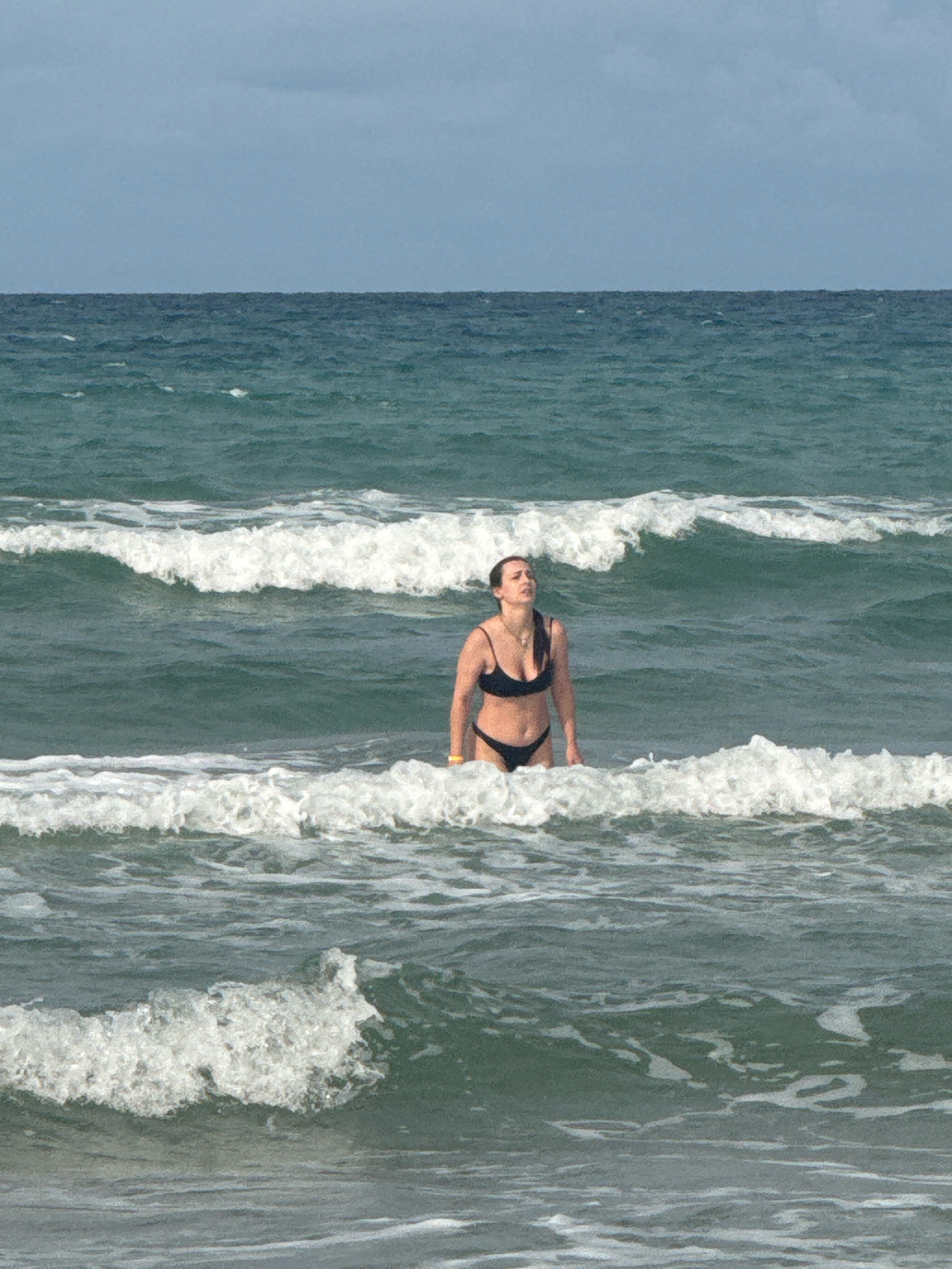 A woman stands in the ocean water at a beach in Anapa, Russia. She is wearing a black bikini. The water is choppy with waves. The sky is overcast.