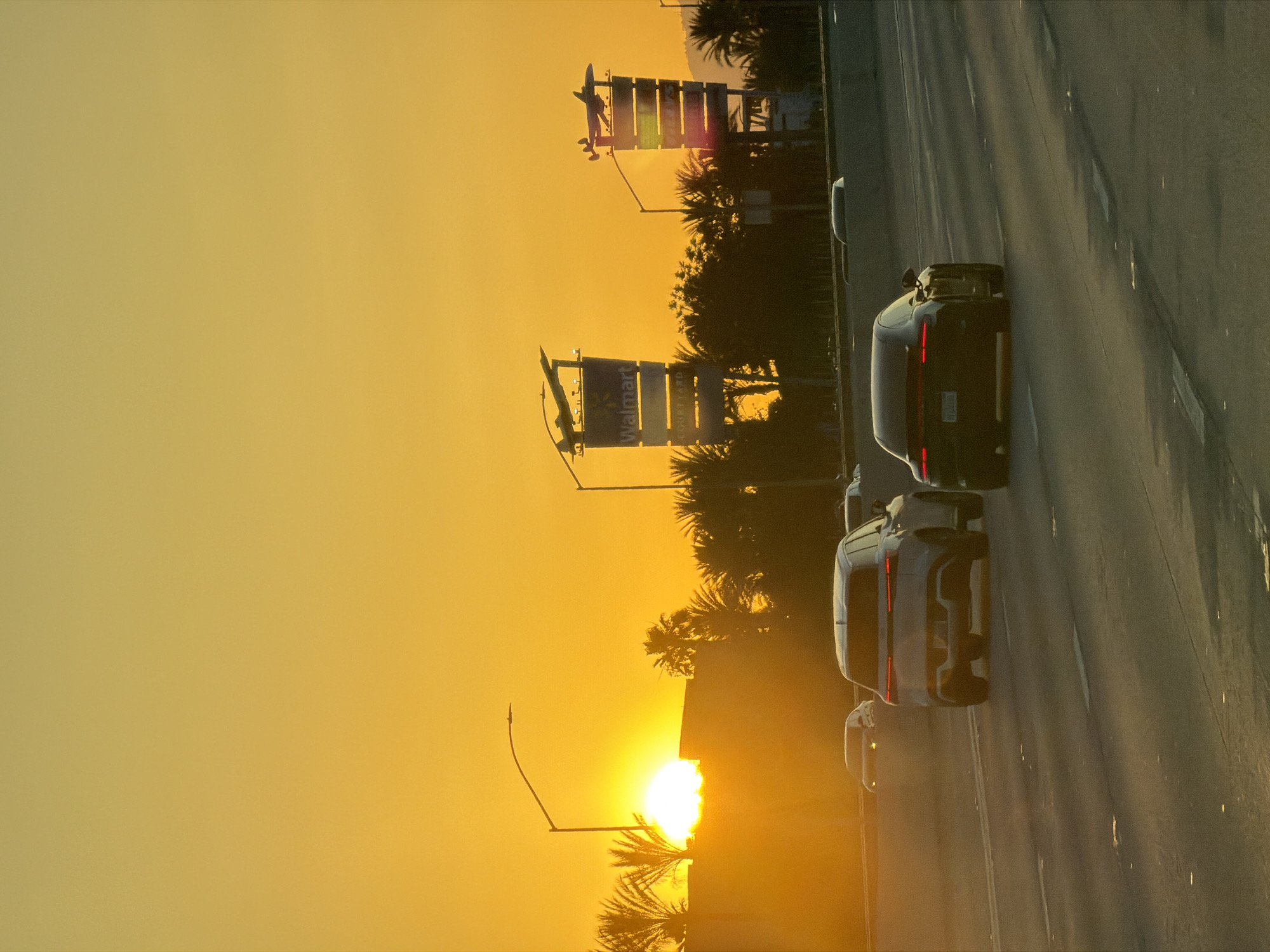 The image shows a sunset scene on a road in Burbank, California, USA. Several cars are driving on the right side of the road, with their rear ends visible. In the background, there are palm trees and the silhouettes of signs for Walmart and Courtyard Marriott, indicating the presence of commercial establishments in the vicinity. The sky is dominated by the setting sun, creating a warm, golden glow throughout the image.