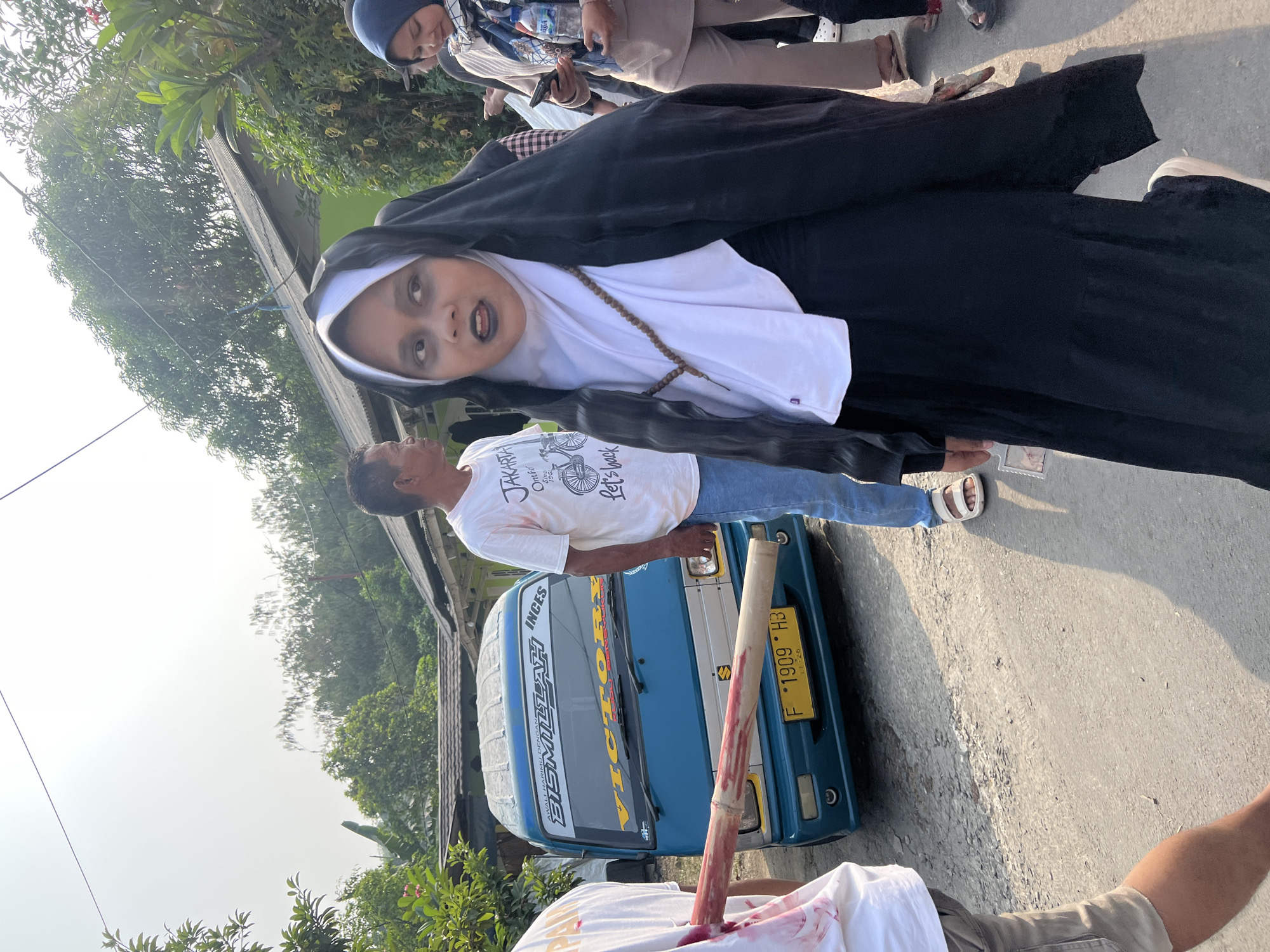 A young person in a nun costume, complete with dark makeup and a rosary, stands in the foreground of a street scene in Klapanunggal, Indonesia. The individual is looking to the right with their mouth open. Behind them, a blue van with "VICTORY" written on the windshield is parked on a gravel road. A person wearing a white t-shirt with "JAKARTA" printed on it stands to the left of the van, and another person in a hijab is visible to the right of the nun costume. The environment is sunny with trees and houses in the background.