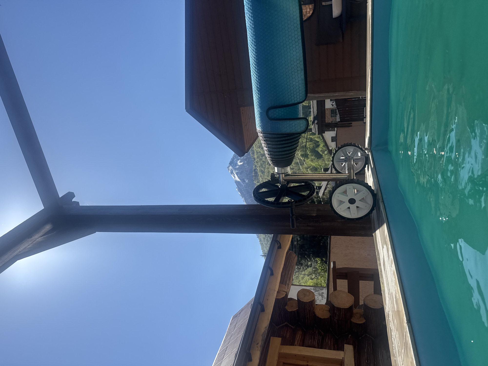The image, taken in Krasnaya Polyana, Russia, shows a partially visible swimming pool with turquoise water. Adjacent to the pool is a rustic, wooden structure decorated with log-shaped accents. Above the pool is a turquoise-colored pool cover rolled up on a wheeled mechanism. In the background, a mountain partially covered in snow is visible under a clear, blue sky. A dark, wooden post and beam structure is in the foreground.