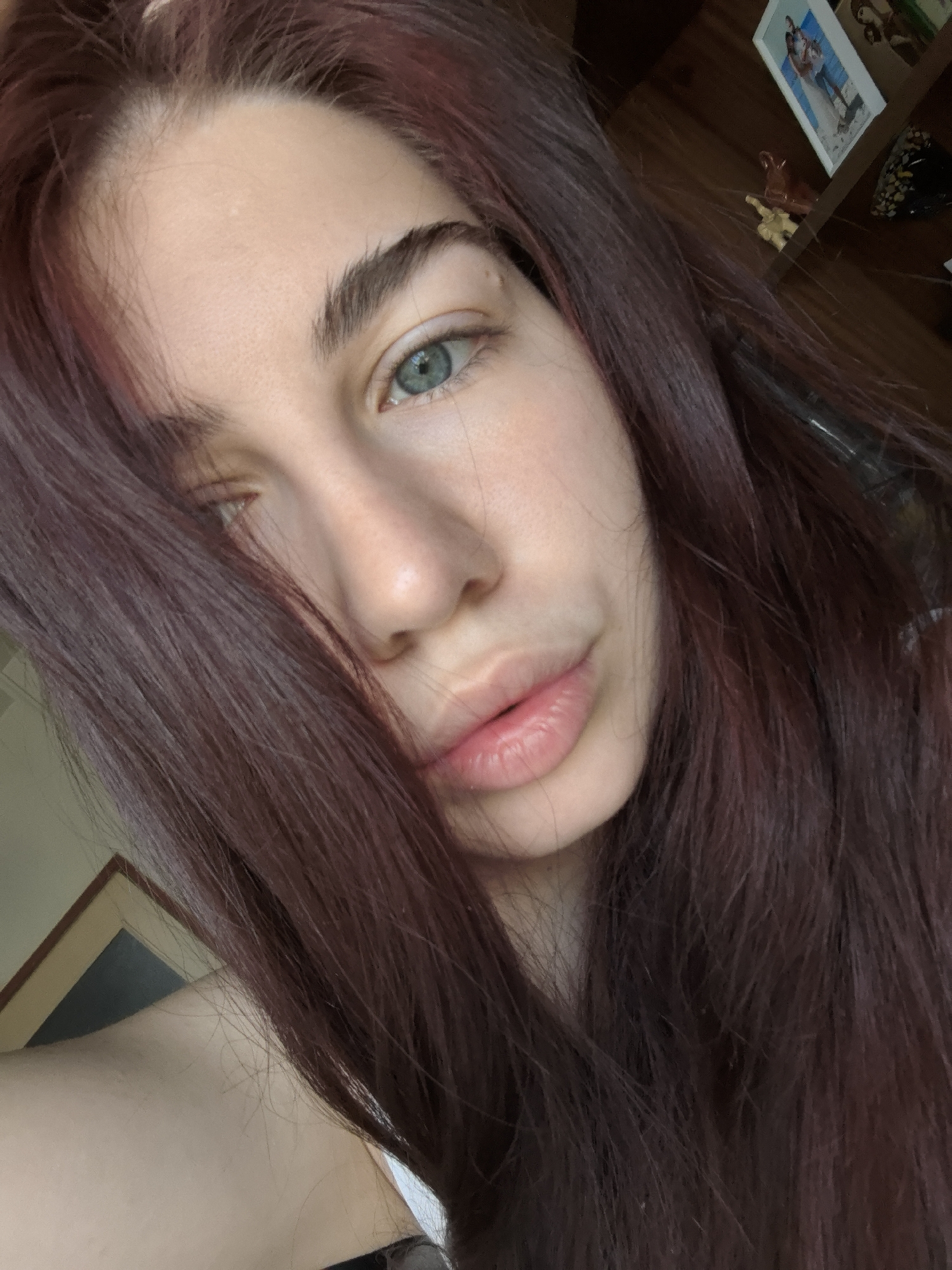 A close-up selfie of a woman with fair skin, light blue eyes, and dark auburn hair that partially covers her face. The photo is taken indoors with a wooden shelf and framed photographs visible in the background, suggesting a domestic setting. The woman's gaze is slightly off-center, and she has a neutral expression. The lighting appears natural. The location in Stara Zagora, Bulgaria is not discernible.