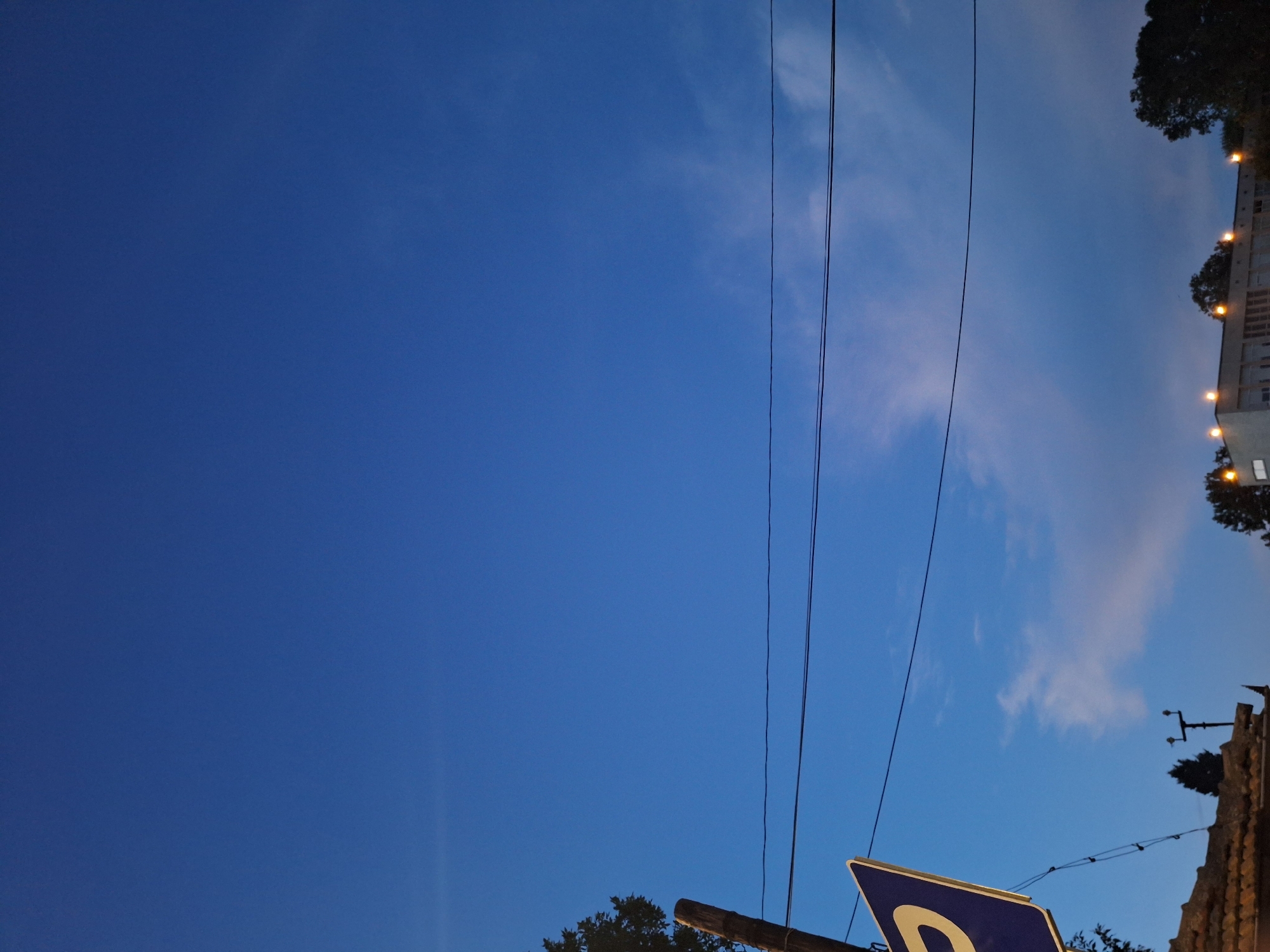 The image captures a scene in Varna, Bulgaria, at dusk. The sky is a deep blue, transitioning from day to night, with faint wisps of clouds visible. In the foreground, a wooden pole supports a blue parking sign with a yellow "P" symbol. Power lines stretch horizontally across the frame, dividing the sky. Below the power lines, the roofline of a building is visible, with several illuminated windows and a few trees silhouetted against the darkening sky. The overall impression is that of an urban landscape settling into evening.