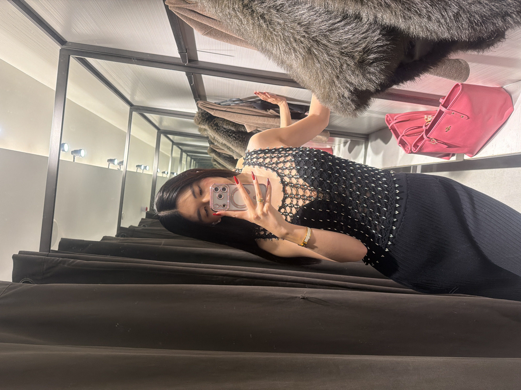 A young woman is taking a selfie in a dressing room, inside a clothing store in what appears to be Paris, France. She is wearing a black top and skirt and is holding her phone up to take the picture. In the background, there are clothes hanging on a rack, including a furry coat, a brown coat, and a pink purse. The dressing room has a gray metal frame and gray curtains. The ceiling is white with rows of small lights. The woman has long, black hair and is wearing red nail polish. She also has a gold bracelet on her wrist.