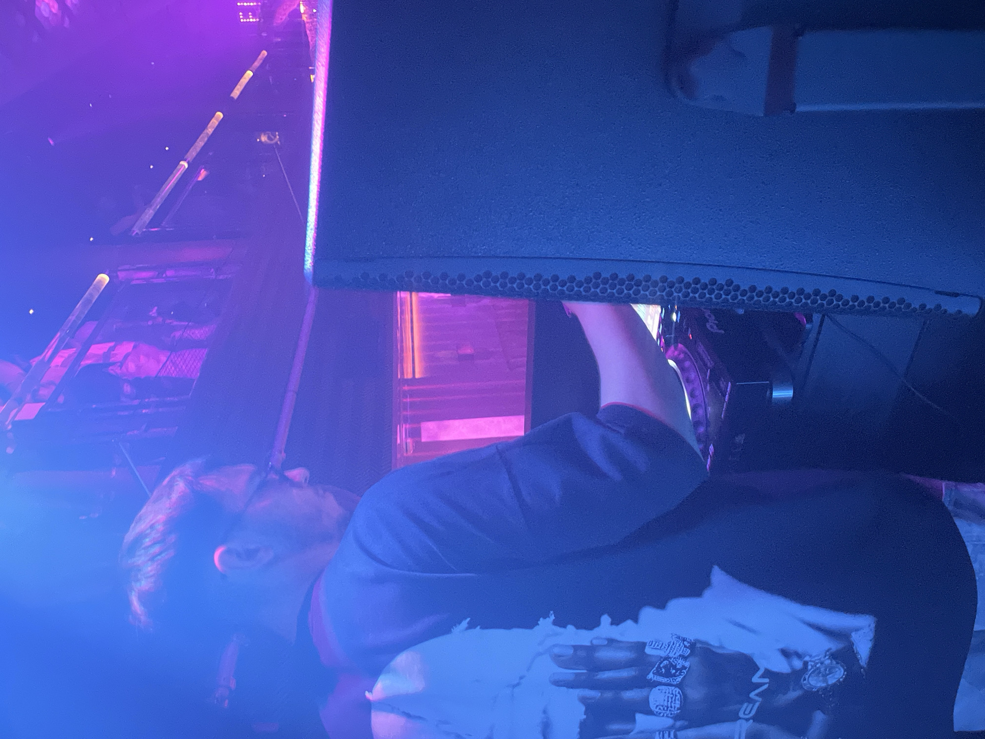 An adult male is observed from the mid-back and right profile, actively engaging with DJ equipment in a dimly lit indoor venue. He possesses short dark hair and wears glasses, a dark short-sleeved t-shirt, and a white t-shirt with a graphic depicting multiple hands wearing rings and the partial word "EAN" is visible on his left shoulder. His right hand is positioned over a black piece of DJ equipment, identified by partial "Pio" branding. A large, dark-colored speaker with a textured, perforated side dominates the immediate right foreground. The environment is illuminated by strong artificial blue and purple lighting. In the background, a multi-level structure or balcony with metal railings is visible, along with multiple horizontal light tubes emitting a warm glow. Vertical illuminated panels with pink light are located behind the DJ setup. The scene depicts a live music performance or club setting in the city of Unknown, Unknown.