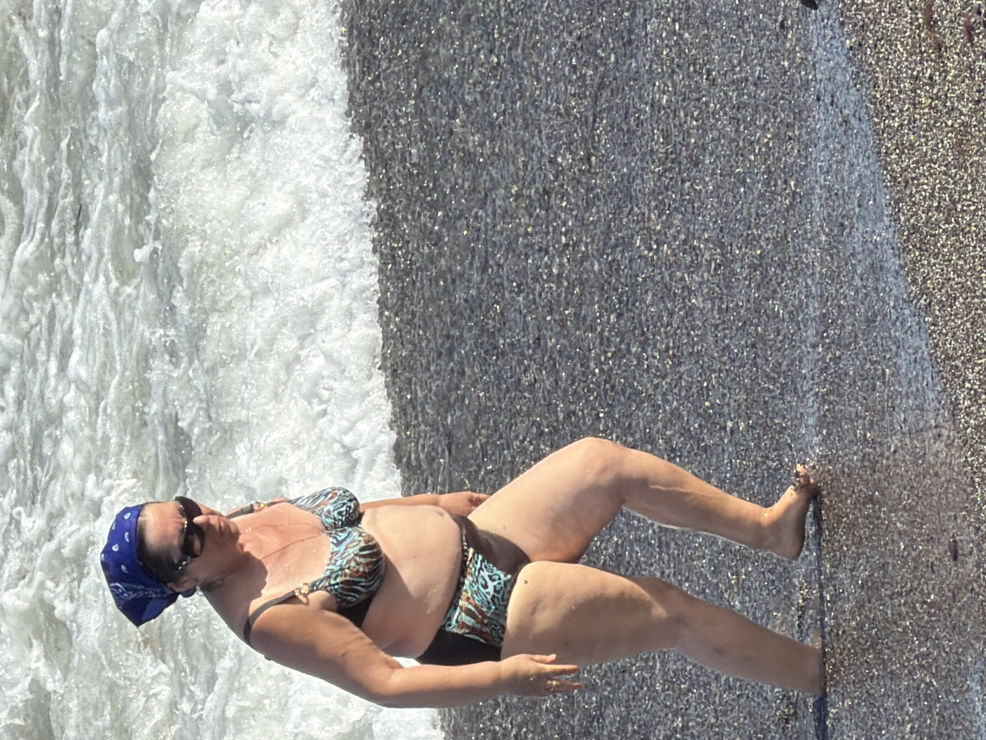 A middle-aged woman wearing a blue bandana, sunglasses, and a patterned bikini walks along the shore of a pebble beach. The waves are breaking near her feet. The water is a light foamy white and turquoise, while the beach is dark grey pebbles. The scene appears to be in Alushta, Ukraine, with the sea in the background.