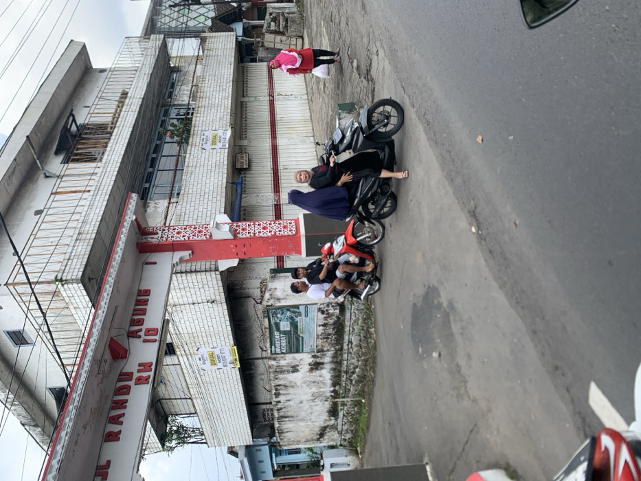 The image depicts a street scene in Singosari, Indonesia (coordinates: -7.874, 112.678). A white building with a red archway inscribed "JL. RAMDU AGUNG 10 RW 10" dominates the left side of the frame. The archway leads to a narrow street with a concrete surface. Three people are visible in the scene. Two are riding a red motorbike in the foreground, and a third person is standing on the sidewalk next to a black motorbike with a female passenger. All three people are wearing casual clothing. There are power lines running along the building, and a sign for "INVESTASI ASIHAT" can be seen on the building's facade. The sky is overcast and visible through a gap in the power lines. The overall atmosphere is quiet and seemingly uneventful.
