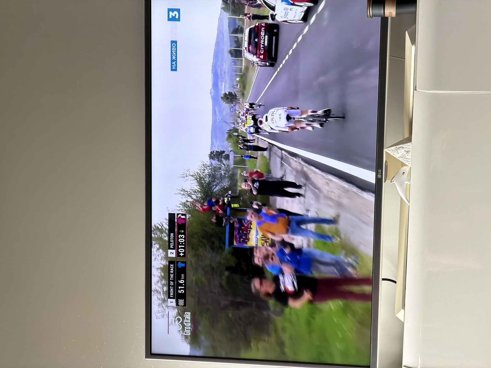 The image shows a television displaying a broadcast of the Giro d'Italia cycling race. The race is passing through a mountainous region with spectators lining the roadside. A Citroen support vehicle is visible in the convoy. On-screen graphics indicate the race information, including that the front of the race is at 51.6 km and the peloton is +01:03 behind. The broadcast channel is "3" with "НА ЖИВО", indicating a live broadcast in Bulgarian. The television is an LG brand. The surroundings suggest it's within a domestic setting. There is no direct evidence within the image to associate it with Plovdiv, Bulgaria.