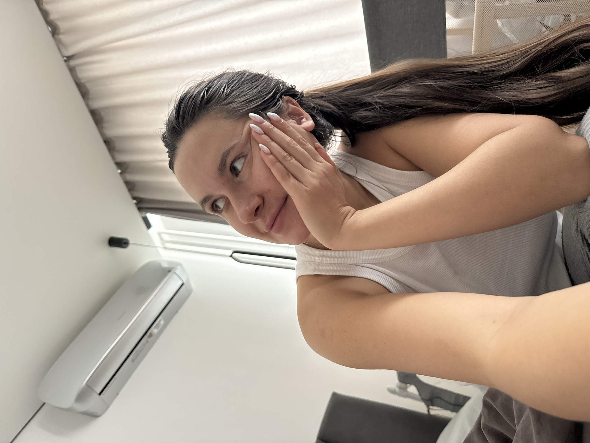 A woman is posing for a selfie in a room with an air conditioner. The room has white walls and a window with white curtains. The woman has long, dark hair and is wearing a white tank top. She is holding her hand up to her face. The location appears to be indoors but the city and specific area of Odintsovo, Russia is not ascertainable from this image.