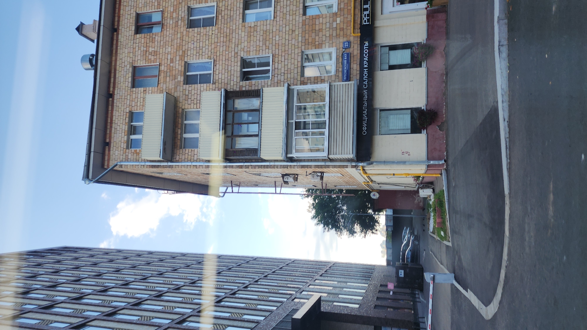 The image shows a street-level view of two buildings and a paved area in Moscow, Russia. On the left is a modern, tall building with a grid of windows. To the right is a brick building with balconies, air conditioning units, and various windows. A sign above a storefront on the brick building reads "ОФИЦИАЛЬНЫЙ САЛОН КРАСОТЫ" (Official Beauty Salon) and also displays "PAUL". Another sign visible is "ПРЕСНЕНСКИЙ ВАЛ 23" (Presnensky Val 23). The ground is paved, and a white and red barrier gate is visible on the left. Several cars are parked in the background. The sky is blue with some clouds.