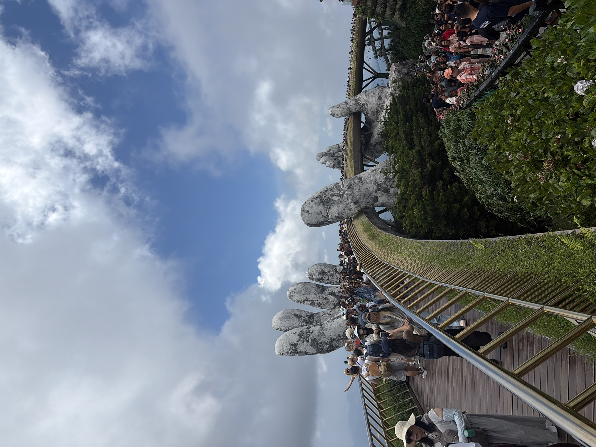 The image depicts the Golden Bridge in Đà Nẵng, Vietnam, a pedestrian bridge supported by two giant, sculpted stone hands. The bridge itself is lined with green foliage. Many people are walking on the bridge. The sky is filled with clouds.