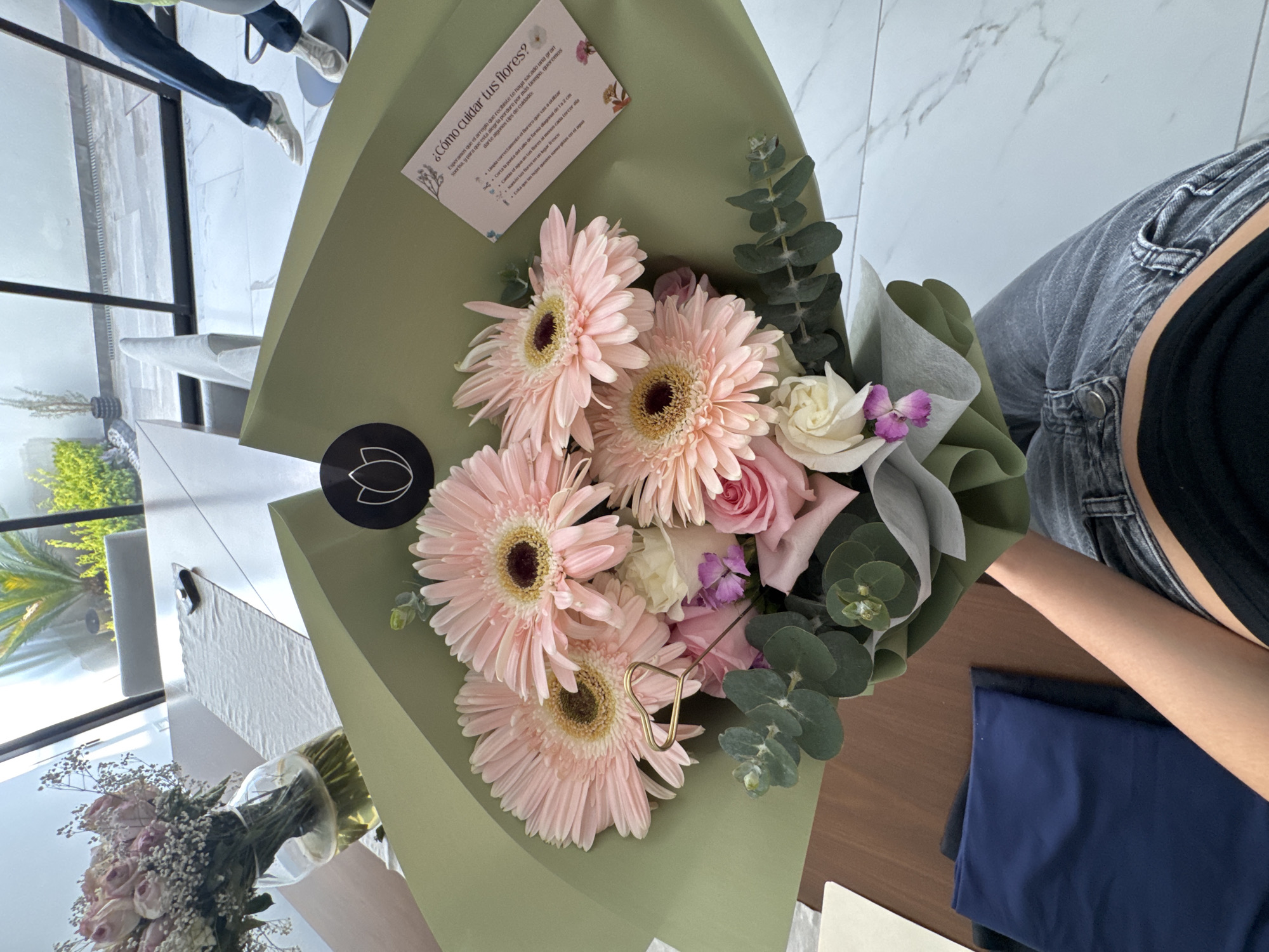 The image depicts a floral arrangement in a green paper wrap, containing gerbera daisies and roses, among other flowers and greenery. A card with care instructions for the flowers is visible in the top left corner. Part of a person's leg wearing jeans and a black top is visible to the right of the arrangement. Another vase of flowers is in the lower-left corner. The setting appears to be indoors, with a view to the outside, possibly in Puebla, Mexico.