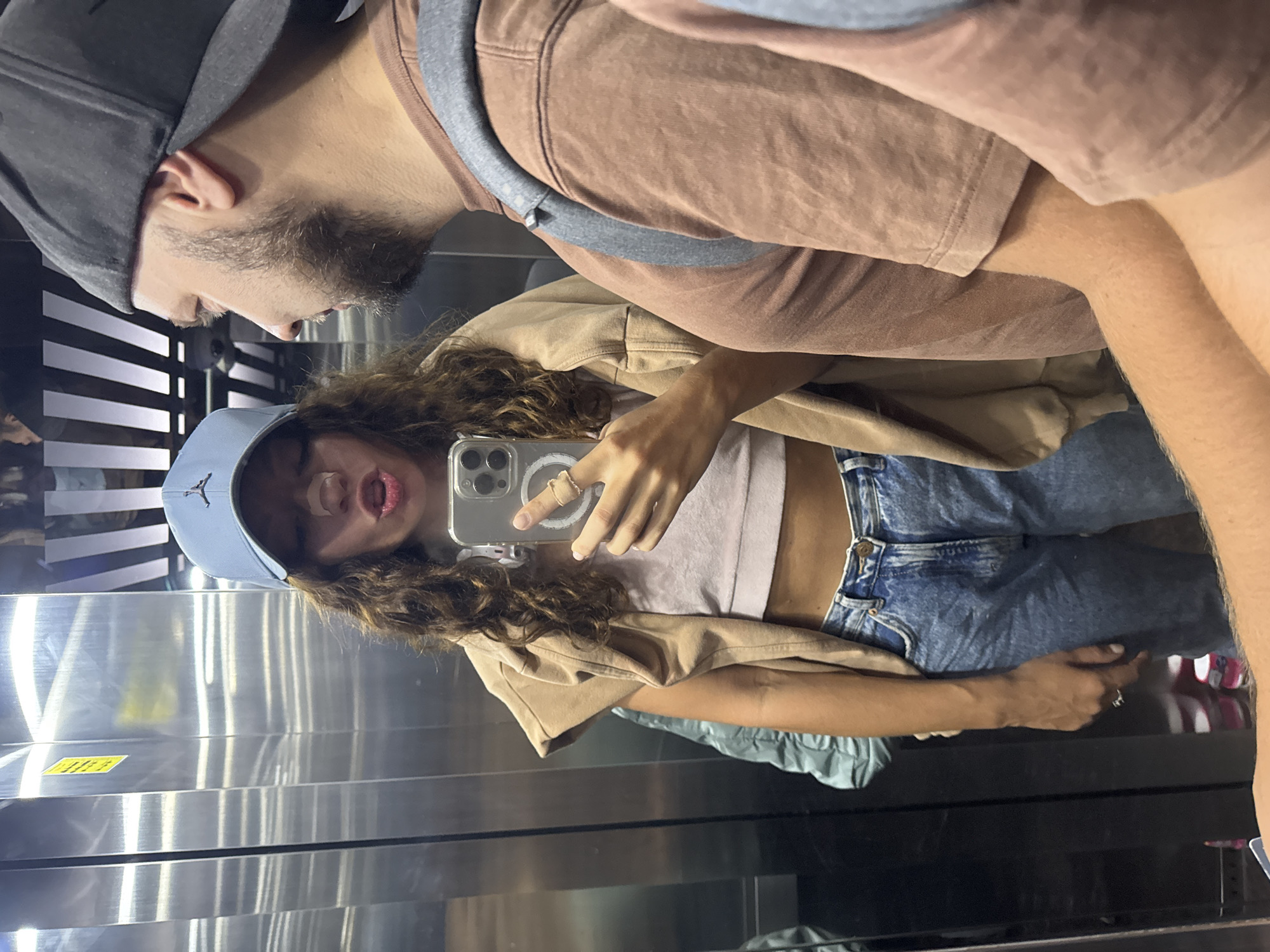 A selfie taken in an elevator. A person with long curly hair wearing a light blue baseball cap, light pink top, tan jacket and blue jeans holds a smartphone. Another person with short dark hair and a dark gray baseball cap is partially visible in the upper left of the image. A person wearing a brown t-shirt is partially visible in the upper center of the image. The elevator is stainless steel with horizontal grooves. The photo's reflection reveals the elevator door's details and hints of the surroundings. There is no information about the city of Đà Nẵng, Vietnam in the image.