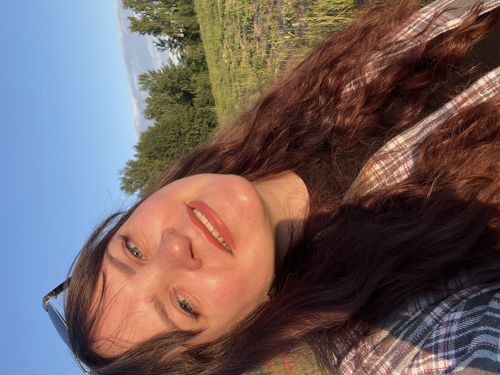 The image is a brightly lit, low-angle selfie of a young woman taken outdoors, depicted as if she is looking up and smiling towards the camera. She has long, wavy, dark brown hair that frames her face, fair skin, and striking light blue or green eyes. Her eyebrows are full and dark, and she wears light reddish-orange lipstick, enhancing her gentle, pleasant smile, which conveys a sense of happiness or contentment. A pair of dark-rimmed sunglasses is partially visible on her head. She is wearing a plaid shirt with a pattern of grey, white, red, and black, visible on her shoulder and chest.
The background shows a natural landscape under a clear blue sky, with a faint, elongated cloud streak. Lush green trees are visible behind her, and beyond them, an open field or grassy area extends into the distance. The lighting suggests late afternoon or early evening, with warm, golden sunlight illuminating her face and hair, creating soft shadows. The overall atmosphere is peaceful and serene. There is no visible text in the image. The context provided indicates the location is Noviy, Russia.