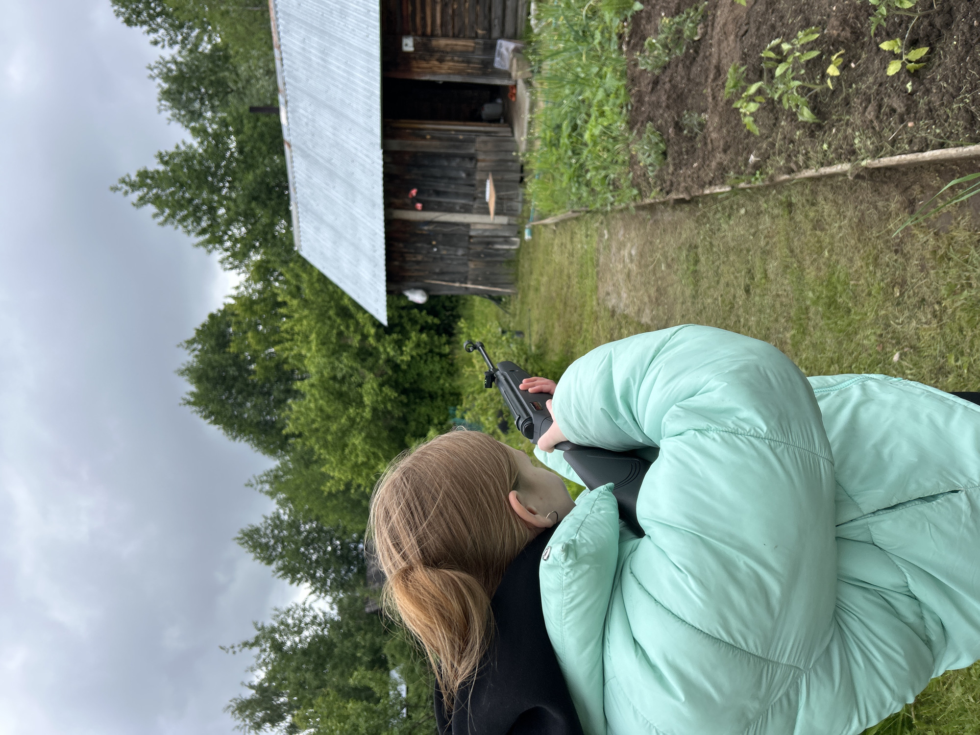 An outdoor scene in the city of Chaikovsky, Russia, depicts a young individual, viewed from behind and slightly to the right, aiming an air rifle. The person has light brown hair tied in a ponytail and is wearing a puffy, light teal/mint green winter or transitional jacket over a black hooded garment. A small silver hoop earring is visible in their left ear. Their body is angled to the left, with the right shoulder shouldering the dark-colored air rifle, which features a fixed front sight and a textured stock.
The weapon is aimed horizontally across a narrow dirt path that divides the foreground. To the left of the path is a strip of mown grass. To the right, a raised garden bed contains dark soil with several small green plants, possibly tomato seedlings, arranged in rows.
In the mid-ground, beyond the garden and path, stands a rustic, dark-stained wooden cabin or shed with a corrugated metal roof. A dark chimney pipe extends from the roof. The cabin's entrance is open, revealing a dark interior and some indeterminate objects. Outside the entrance, a small, light-colored wooden table is positioned. Various items, including what appears to be a white bag and a long stick, are leaning against the cabin's exterior walls.
The background consists of dense green foliage, primarily trees, under an overcast, grey sky. The overall light suggests a cloudy day.