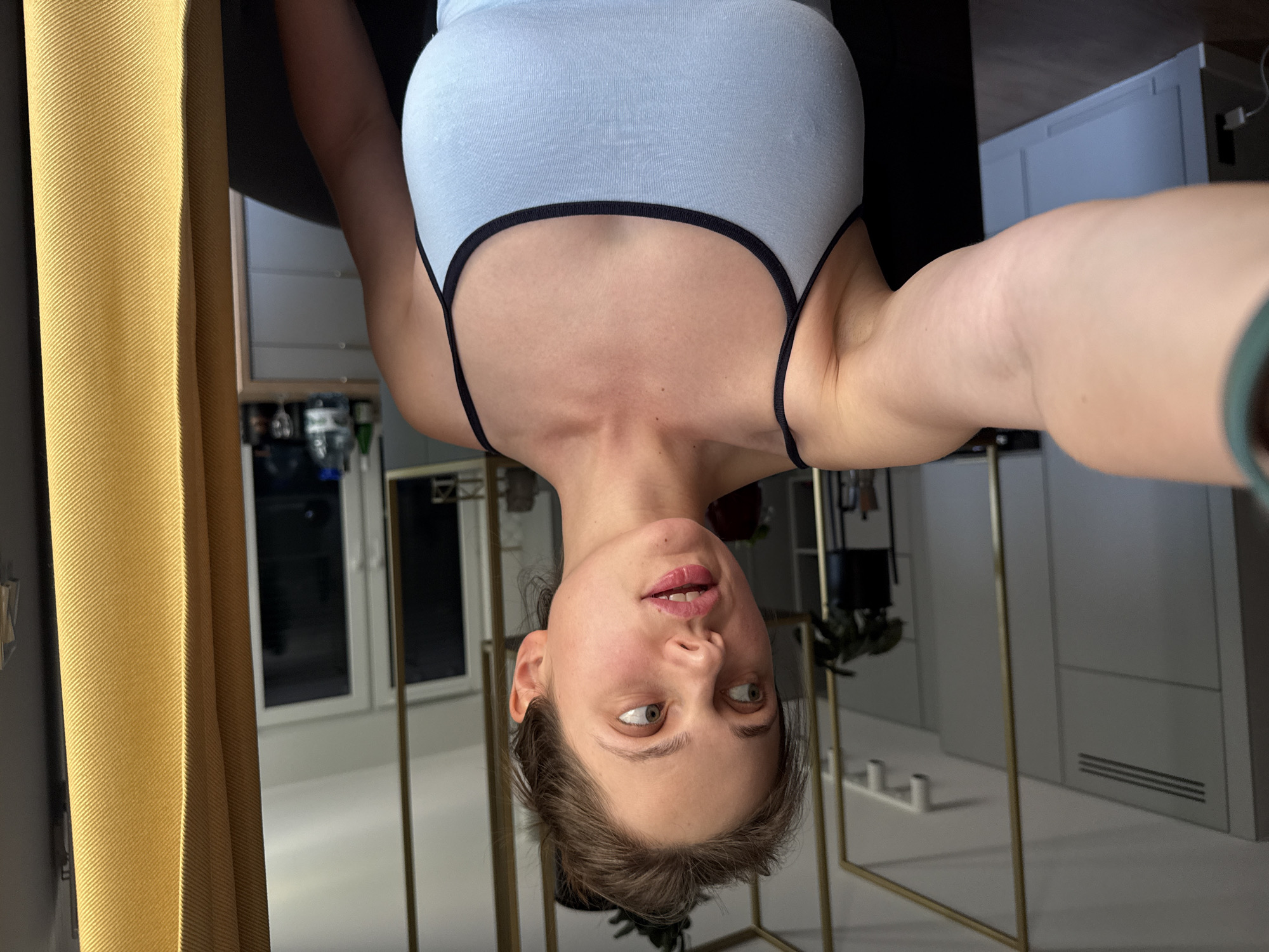 A woman is captured in a selfie, her head oriented downwards, suggesting she is lying on a surface or hanging upside down. She wears a light blue tank top with black trim. Her facial expression is playful and curious, with wide eyes looking to her right and slightly parted lips. The background shows an interior setting with modern elements. To the left, a textured yellow curtain is visible, and to the right, there are white cabinets and a sleek, minimalist design. A metal structure, possibly a mirror frame or furniture, is also present. The lighting appears to be indoor, suggesting a daytime or evening scene within a home or studio. There is no visible text.
