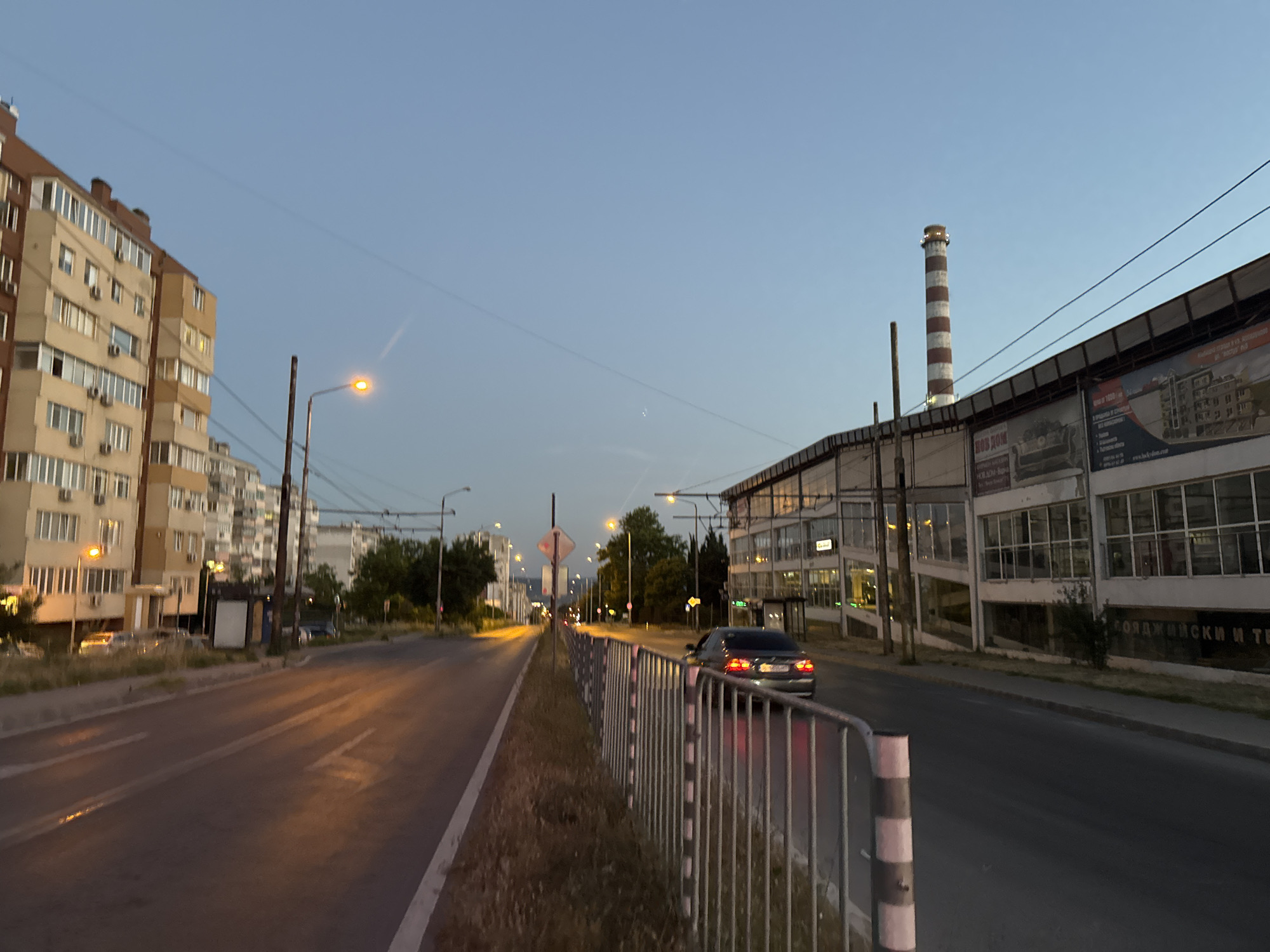 The image depicts a street scene in Varna, Bulgaria, during twilight. The perspective is from the side of a road, looking forward. On the left, a multi-story residential building with light-colored facades and numerous balconies stretches into the distance. Streetlights with orange bulbs are visible, illuminating sections of the road and sidewalk. On the right side of the road, there is a long, modern building with a glass facade, behind which a tall, striped industrial chimney with red and white bands is prominent against the pale blue sky. A dark-colored sedan is driving away from the camera on the right lane of the road. A metal fence runs along the right side of the sidewalk, separating it from the road. The sky is clear with a faint contrail visible. The road surface shows reflections from the streetlights.