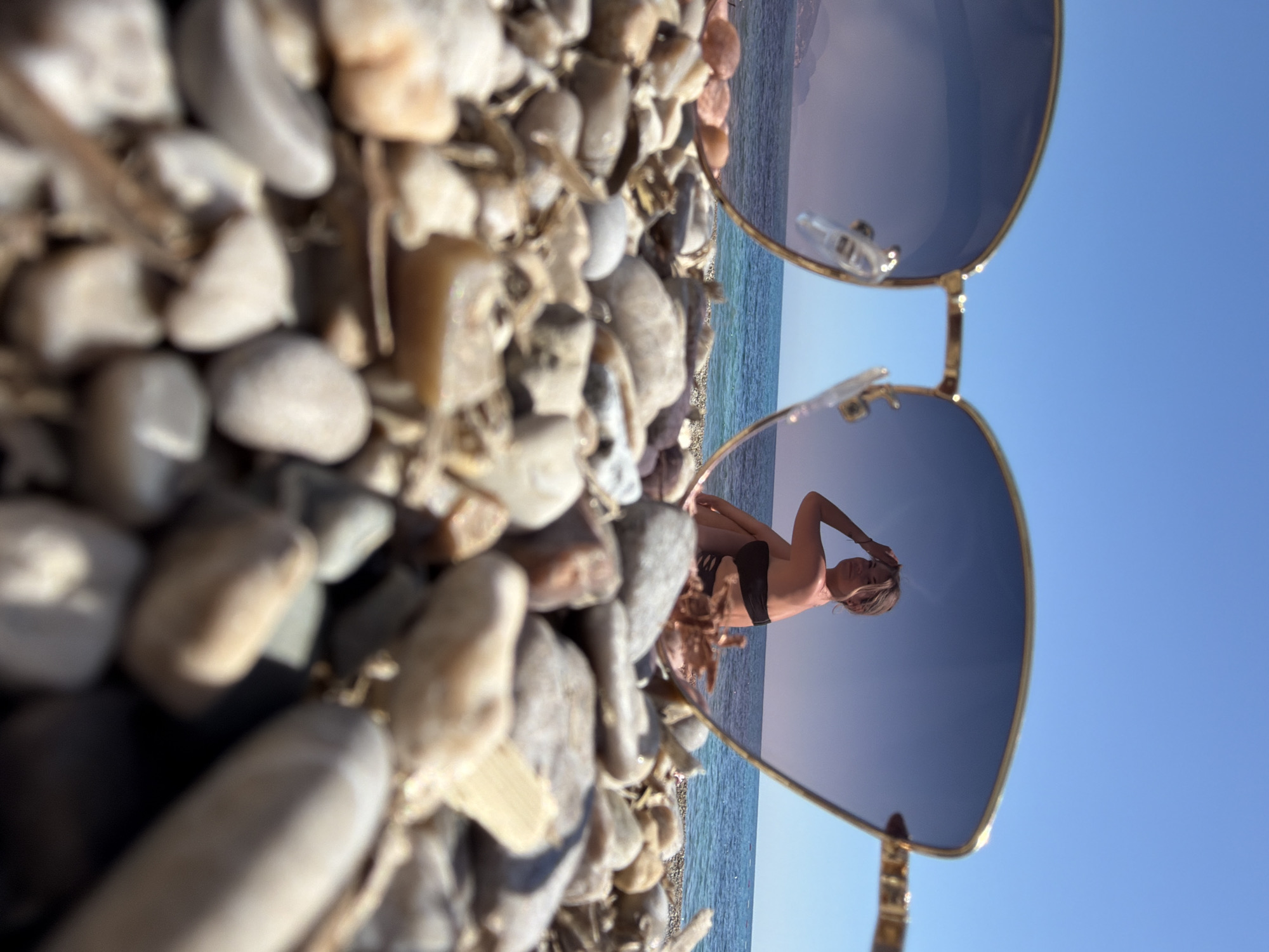 The image depicts a beach scene with pebbles in the foreground, and a pair of sunglasses in the center frame. The reflection within the sunglasses shows a person, likely a woman, posing in a black swimsuit against a backdrop of the sea and clear blue sky. The location is believed to be Prijevor, Montenegro, given the typical Mediterranean coastline and pebble beaches found in that region. The pebbles vary in size, shape, and color, mainly shades of grey and brown. The sunglasses have gold frames. The person in the reflection appears to be standing on the beach near the water, with one hand raised towards their head. The sea appears calm and blue, blending into the cloudless sky.