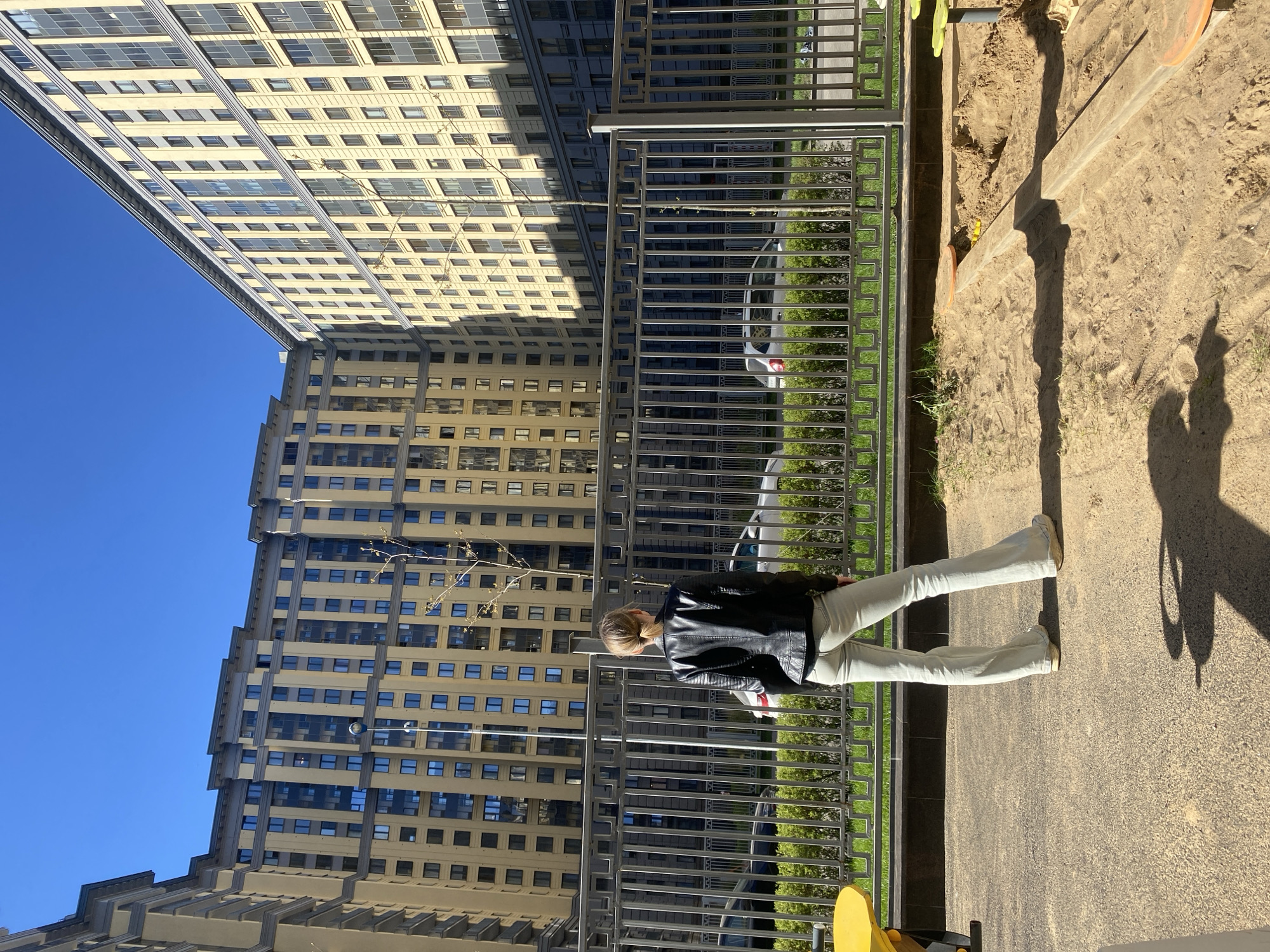A woman wearing a black leather jacket and light-colored jeans stands next to a metal fence. On the other side of the fence is a green space with trees and a parked car. To the left of the fence, there is a tall, beige-colored building with numerous windows under a clear blue sky. The shadow of the woman and the fence can be seen on the ground to the right. The image appears to be taken outdoors in an urban setting, possibly in Saint Petersburg, Russia given the architectural style of the building.