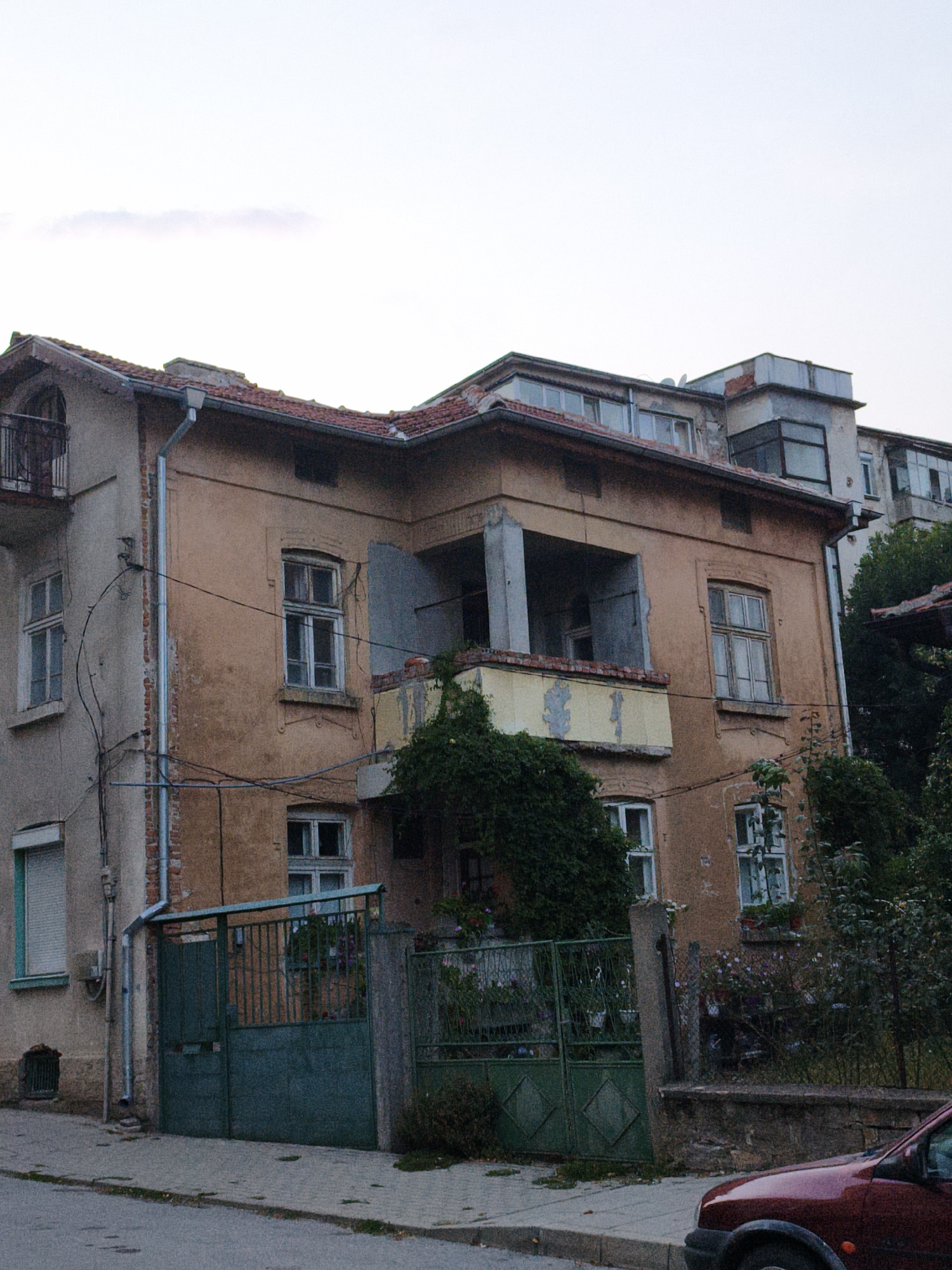 The image captures a view of an old, two-story building with peeling, ochre-colored paint, located in the city of Lovech, Bulgaria. The building features multiple windows, some with white frames and shutters, and a balcony on the second floor. Overgrown green ivy climbs the building's facade and covers parts of the balcony. A green metal gate stands in front of the building, partially obscuring a courtyard with more greenery. A modern red car is parked on the street in the foreground, to the right of the gate. The sky is overcast, suggesting a cloudy day.
