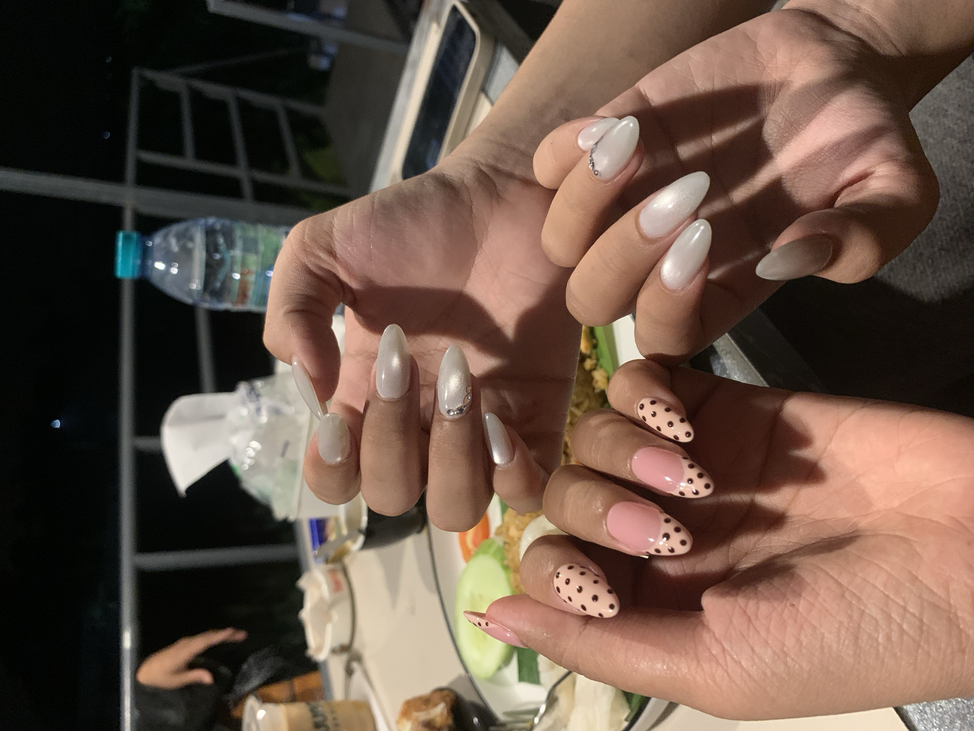 This image captures a close-up view of two pairs of hands adorned with intricately designed artificial nails, presented on a table setting. The background suggests an outdoor dining environment, possibly a restaurant or cafe, with railings and ambient lighting indicating it's nighttime. In the background, out of focus, there's a table with food, a bottle of water, and a napkin dispenser, implying a meal in progress. The overall scene suggests a social gathering, likely a dinner outing, in Jajag, Indonesia.