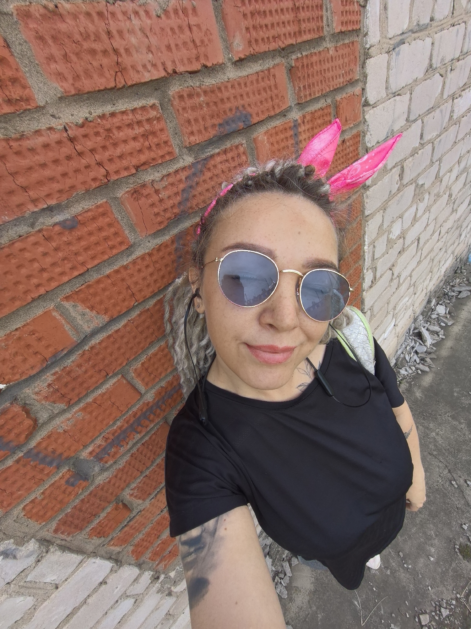 A woman takes a selfie outdoors, positioned between two brick walls, one red and the other white. She wears round sunglasses, a black shirt, and has braided hair with pink bunny ears. The location is Noviy, Russia. The ground is concrete with scattered debris.