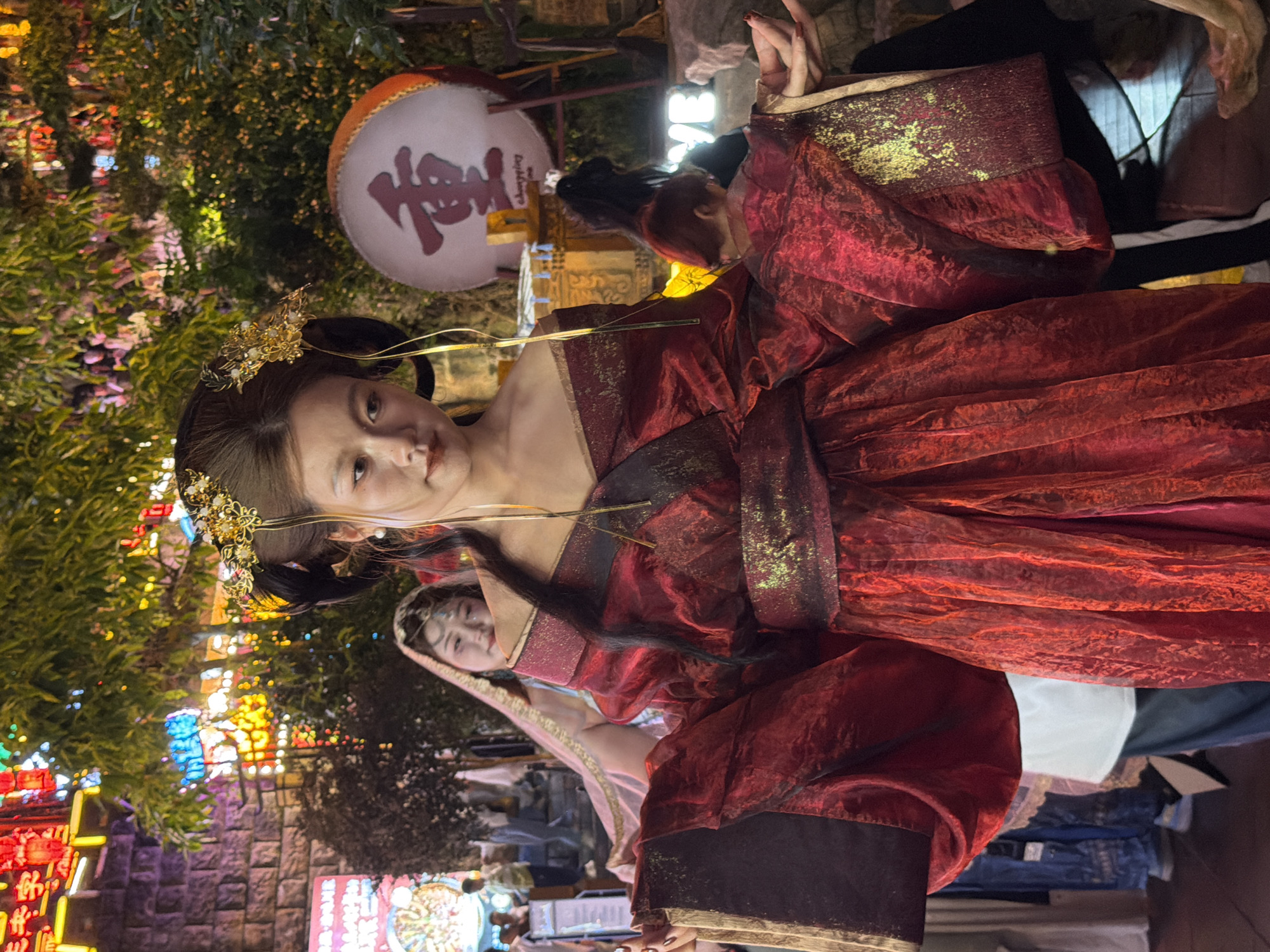A young East Asian woman stands in the foreground, wearing a red, off-the-shoulder traditional Chinese garment adorned with gold accents. Her hair is styled in an elaborate updo with golden hair ornaments. She is looking slightly down and to the right, with her arms extended outwards. Behind her, another woman in a veil and lighter colored clothing looks towards the camera. In the background, there are colorful neon lights and foliage. A large drum with Chinese characters on it is visible to the right of the woman in the foreground. The scene appears to be outdoors at night, likely in an entertainment district, possibly in Yuzhong District, China, given the presence of traditional Chinese attire and signage.